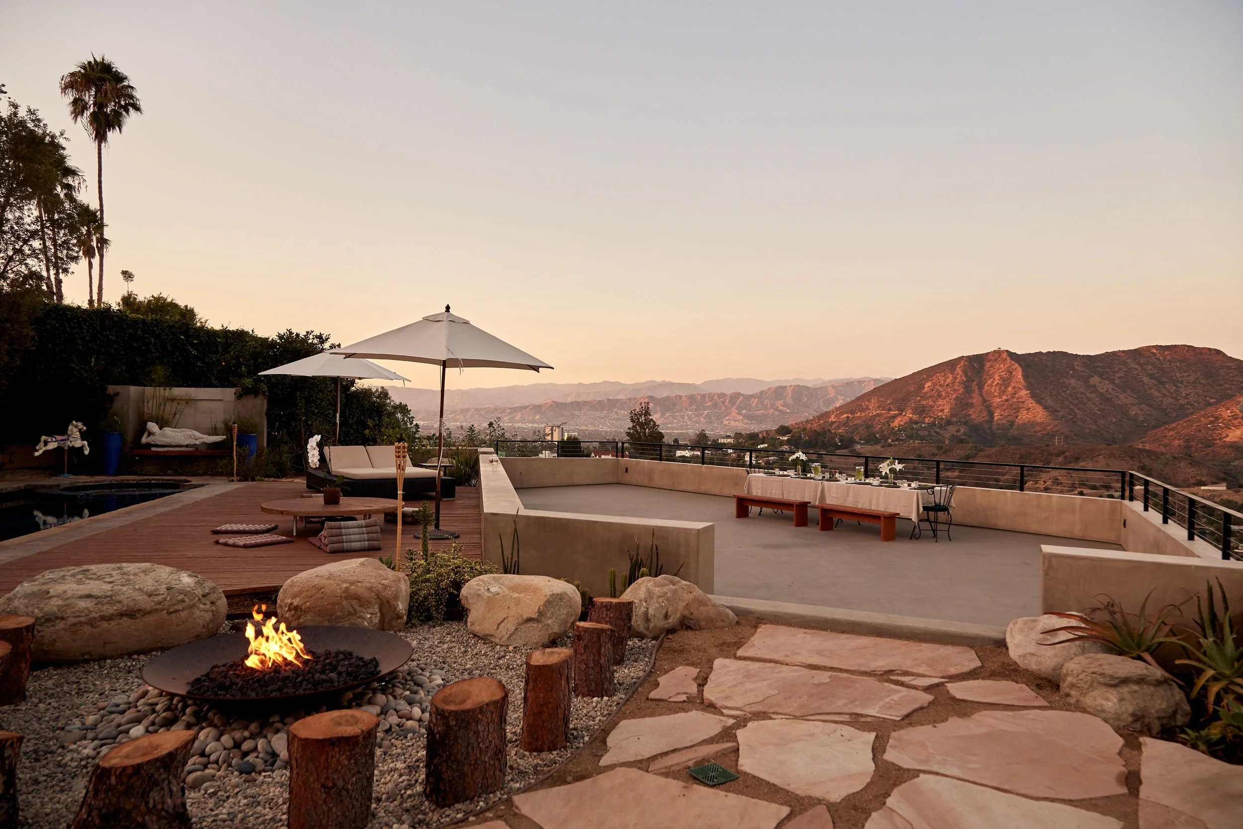 Outdoor patio with a fire pit, deck chairs, and umbrellas overlooking a scenic mountain view at sunset.
