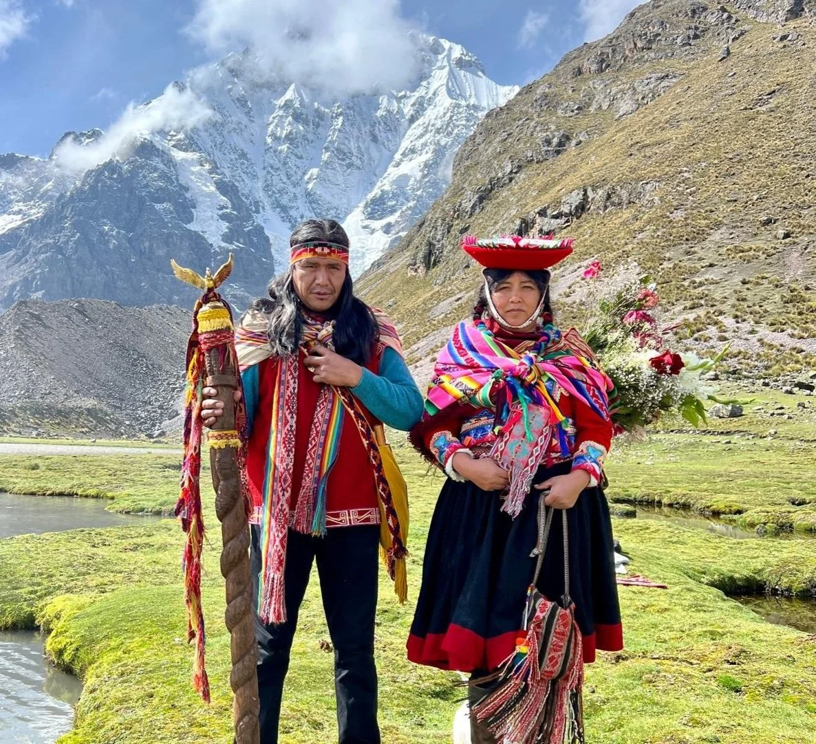 Two people in traditional Andean clothing standing in a mountainous landscape with snow-capped peaks in the background.