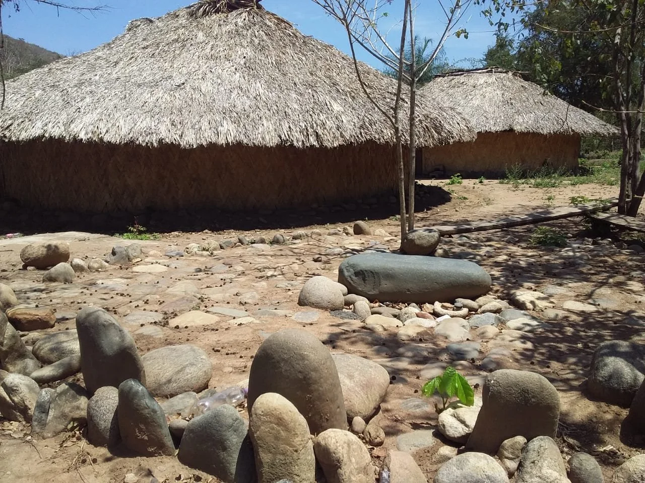Traditional thatched roof huts surrounded by rocks and stones, with sparse vegetation and trees under a sunny sky.