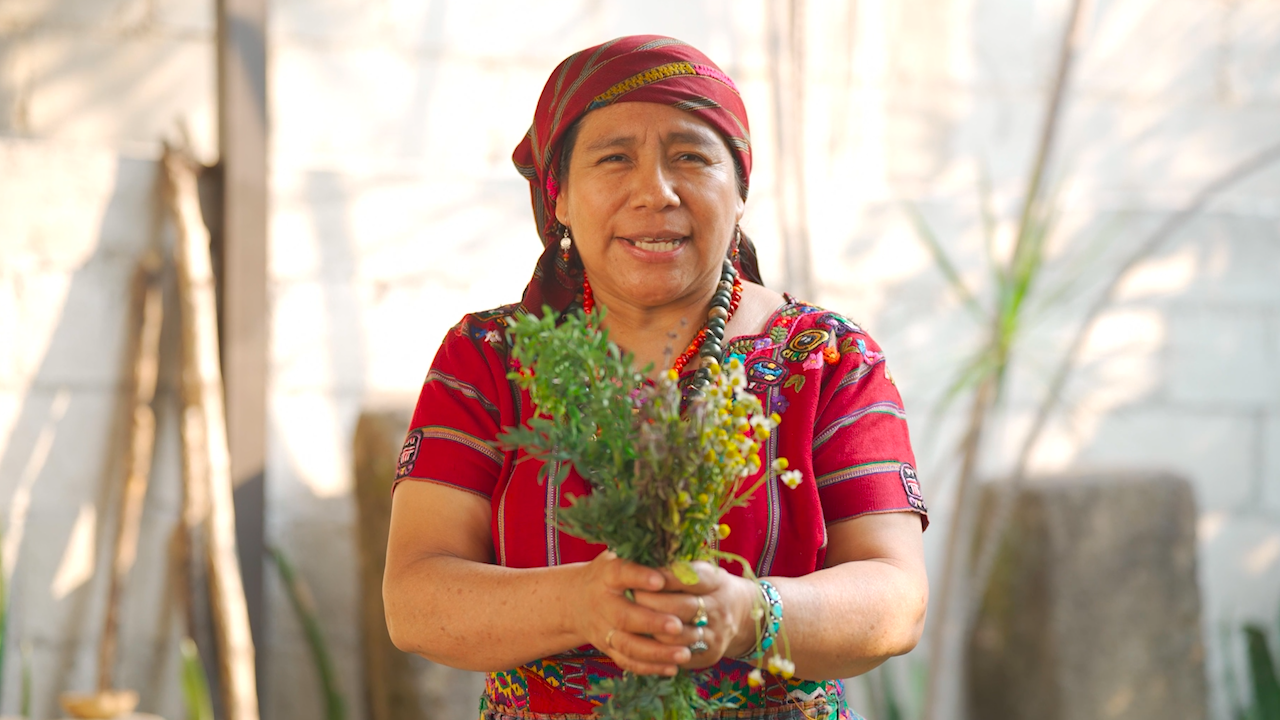 A man wearing a colorful, traditional-looking garment and a vibrant headscarf, sitting outdoors in front of a tree with green foliage.