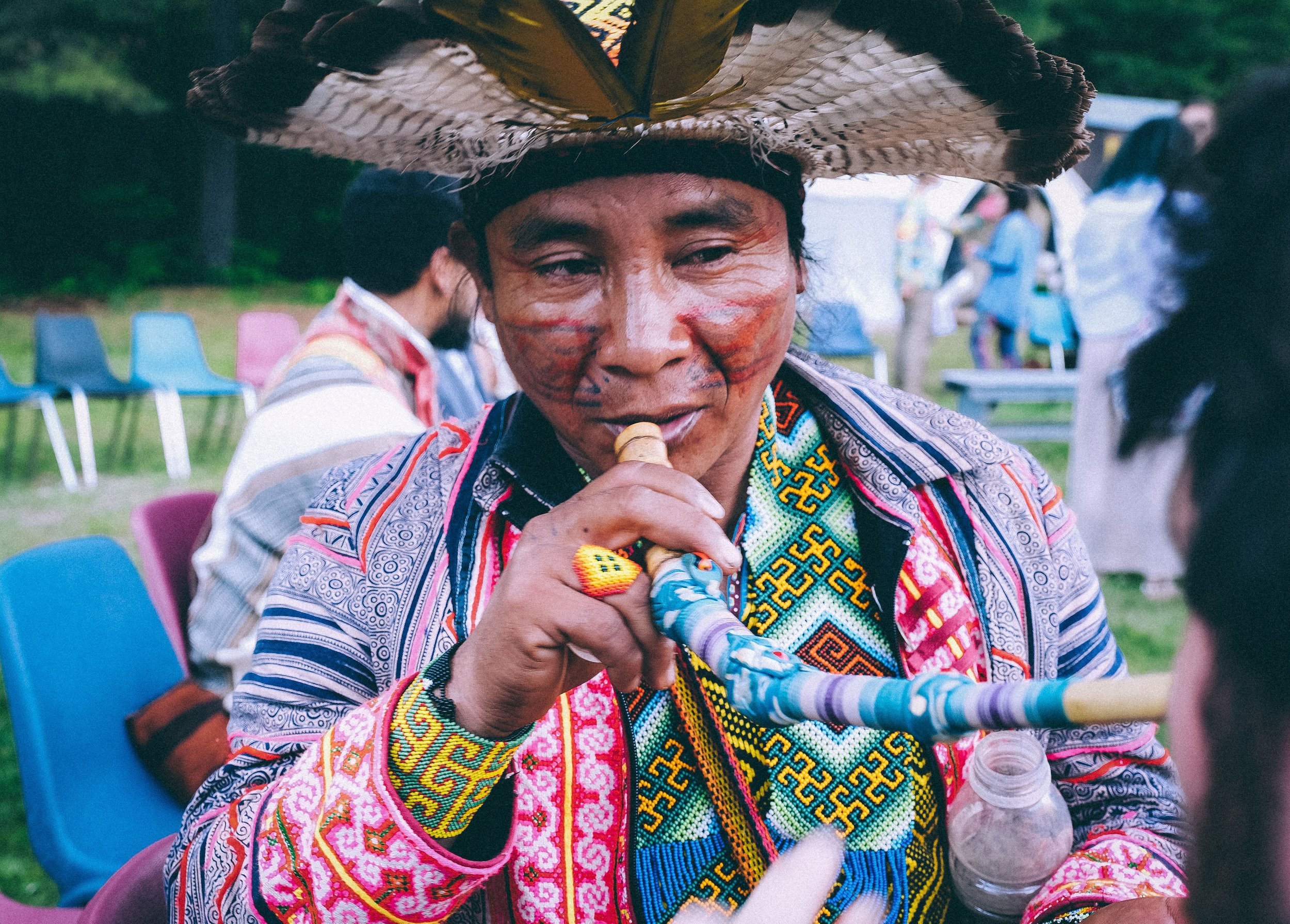A woman with facial paint and traditional colorful clothing, wearing a large woven hat, drinking from a bottle at an outdoor gathering with colorful chairs and other people in the background.
