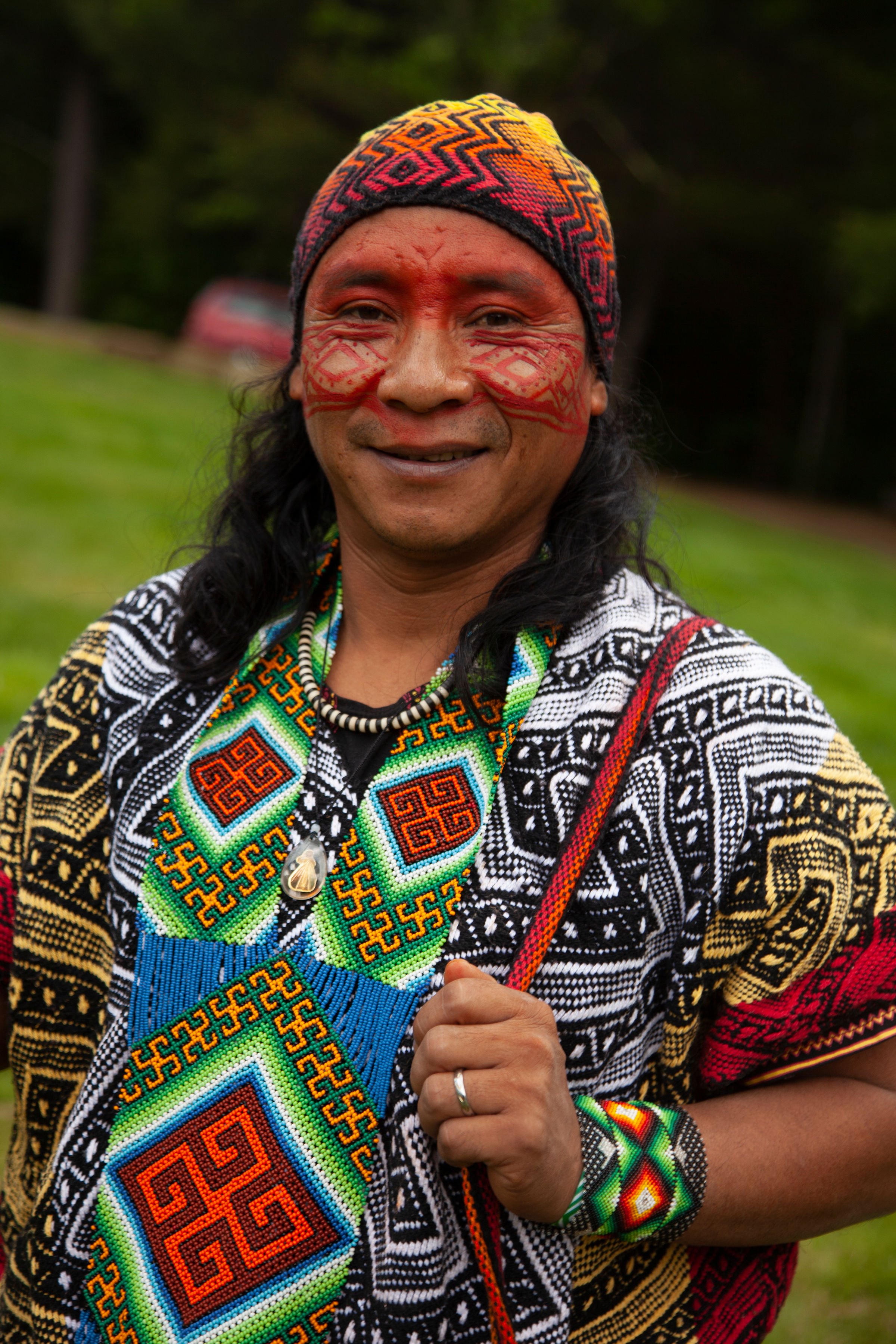 A man with long black hair, wearing traditional clothing and face paint, smiling outdoors in a green setting.