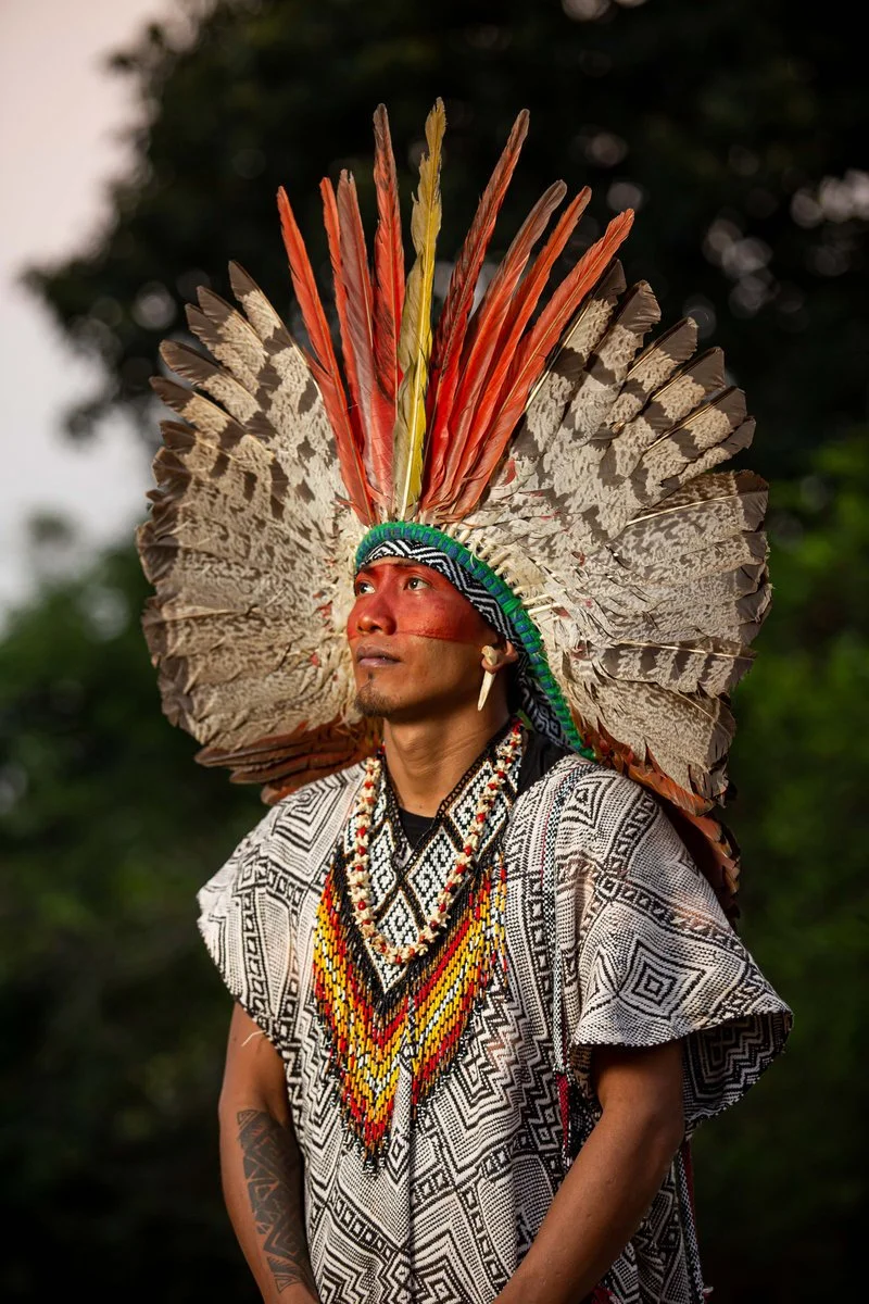 A man wearing traditional indigenous attire, including an elaborate feather headdress with red, brown, and beige feathers, and a beaded necklace with bright yellow, red, and black beads. He has a face paint stripe across his face and an earring, standing outdoors with blurred green trees in the background.