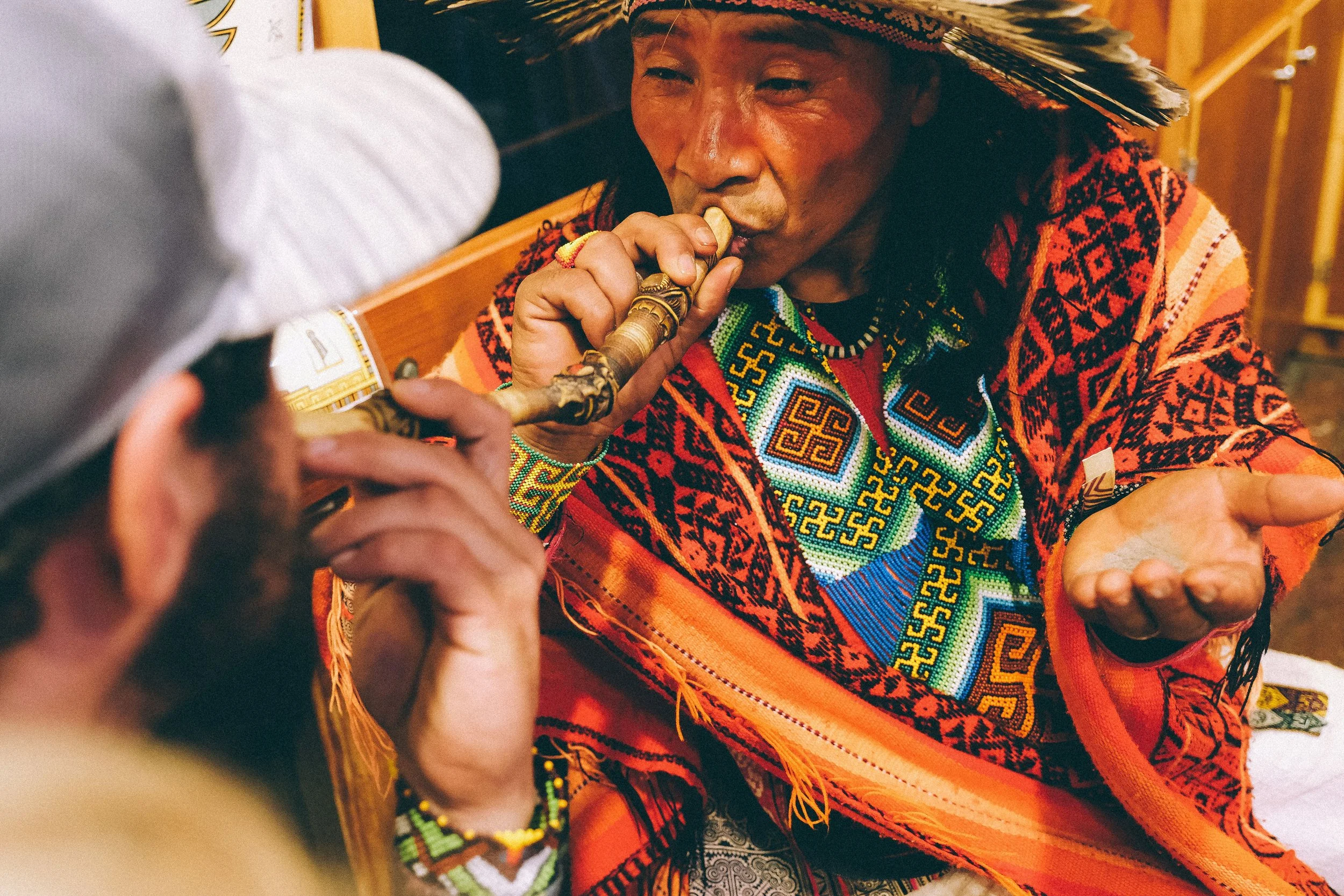 A person wearing traditional colorful, beaded clothing and a feathered headdress plays a wind instrument in a cultural setting.