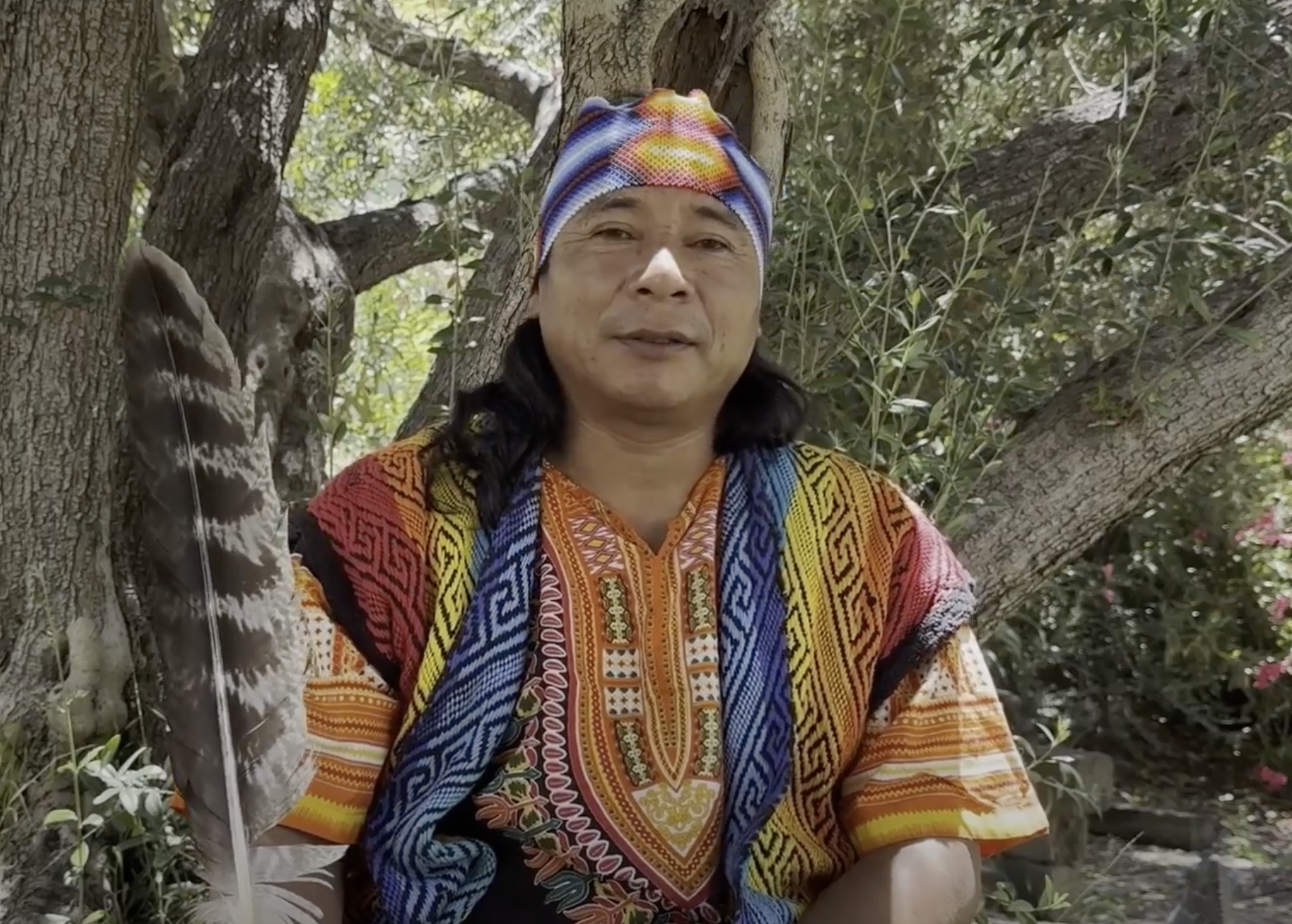 A man wearing a colorful, traditional-looking garment and a vibrant headscarf, sitting outdoors in front of a tree with green foliage.