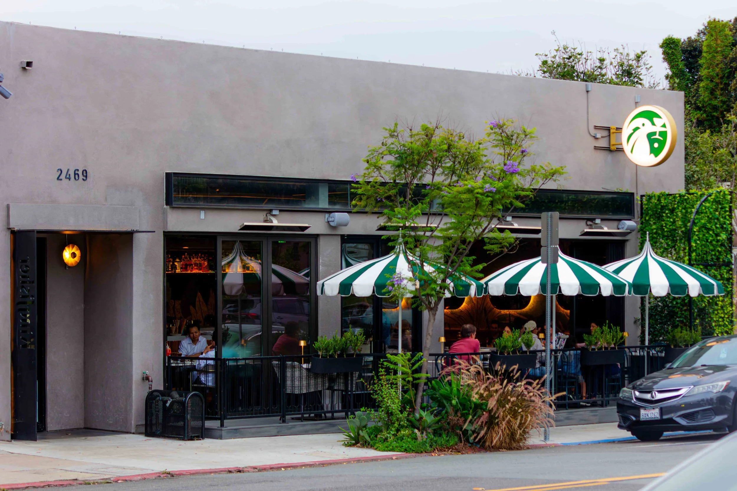 Exterior view of a restaurant with seating area on a sidewalk, striped green and white umbrellas, a few people dining outside, trees, and a city street with cars parked along the curb.