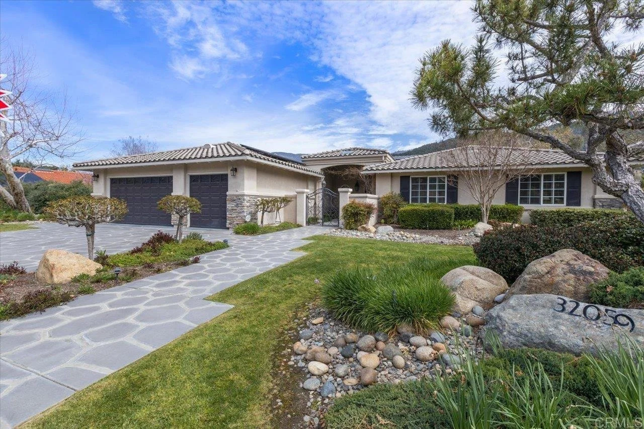 Front yard of a single-story house with a paved walkway, well-maintained grass, trees, shrubs, and rocks. The house has a beige exterior, black shutters, and a double garage with black doors. House number 32059 is visible on a large rock.