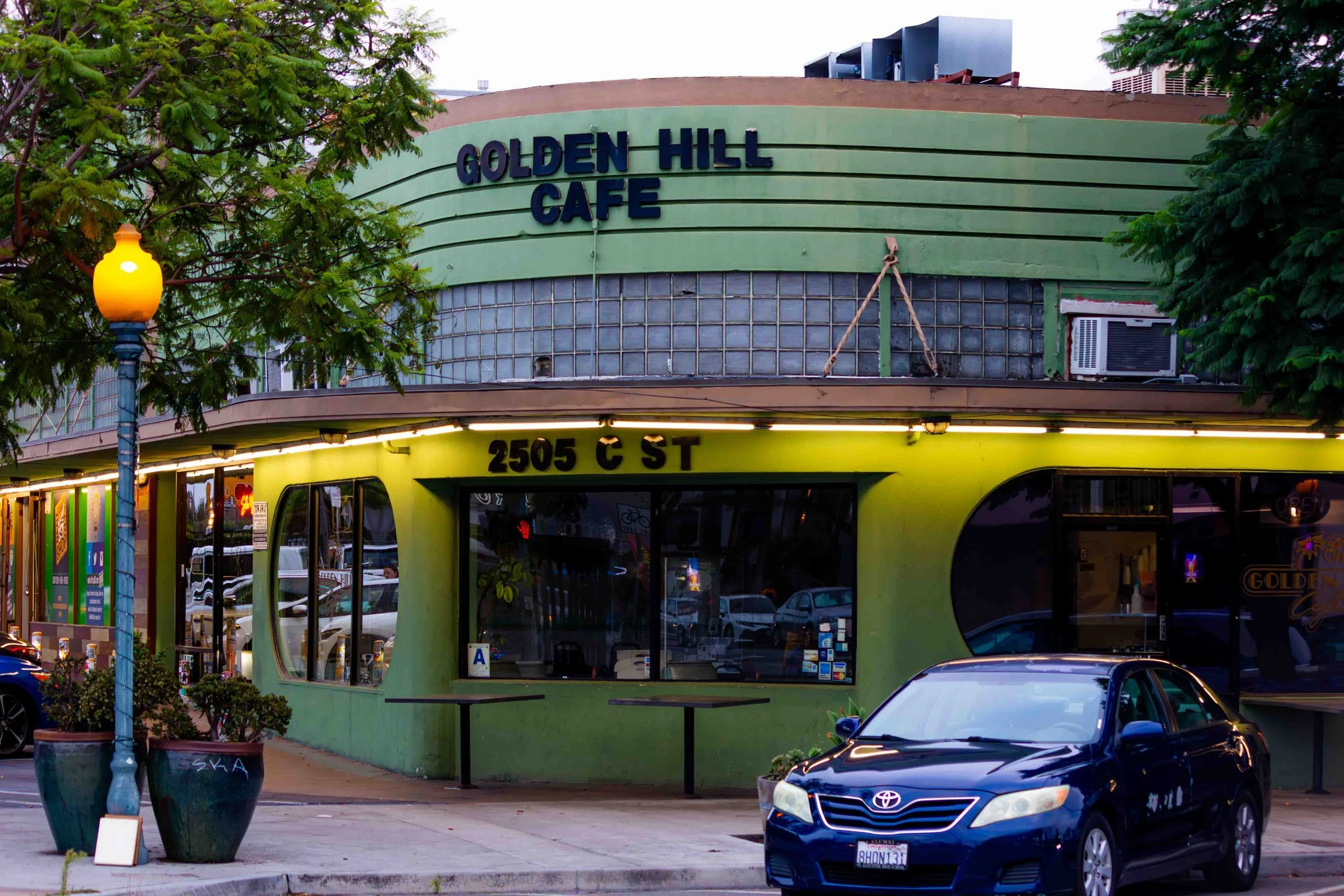 The image shows a green building with a sign that says 'GOLDEN HIll CAFE' on the upper part. The lower part of the building has large windows and a yellow awning with the address '2505 C ST.' There is a black car parked in front and a street lamp on 
