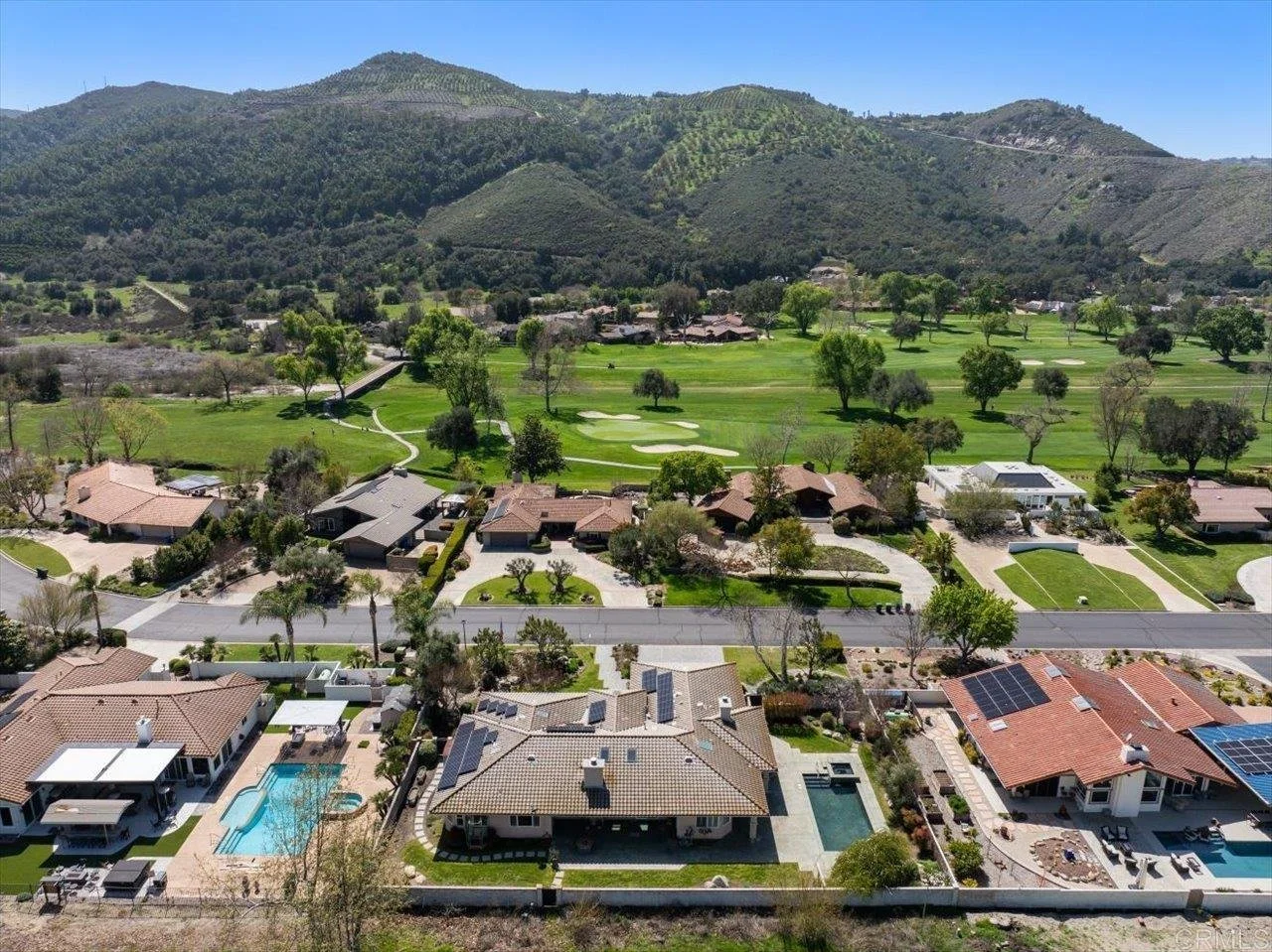 Aerial view of a residential neighborhood with houses, pools, and green lawns, adjacent to a golf course with trees and sand traps, set against rolling hills in the background.