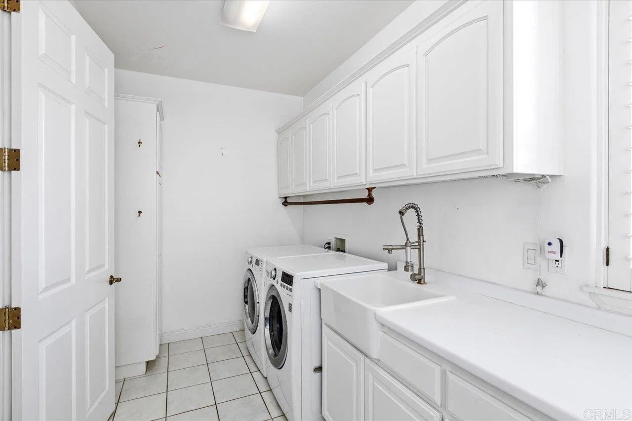 A laundry room with white cabinets, a sink, a front-loading washing machine and dryer, a window with blinds, and tiled floor.