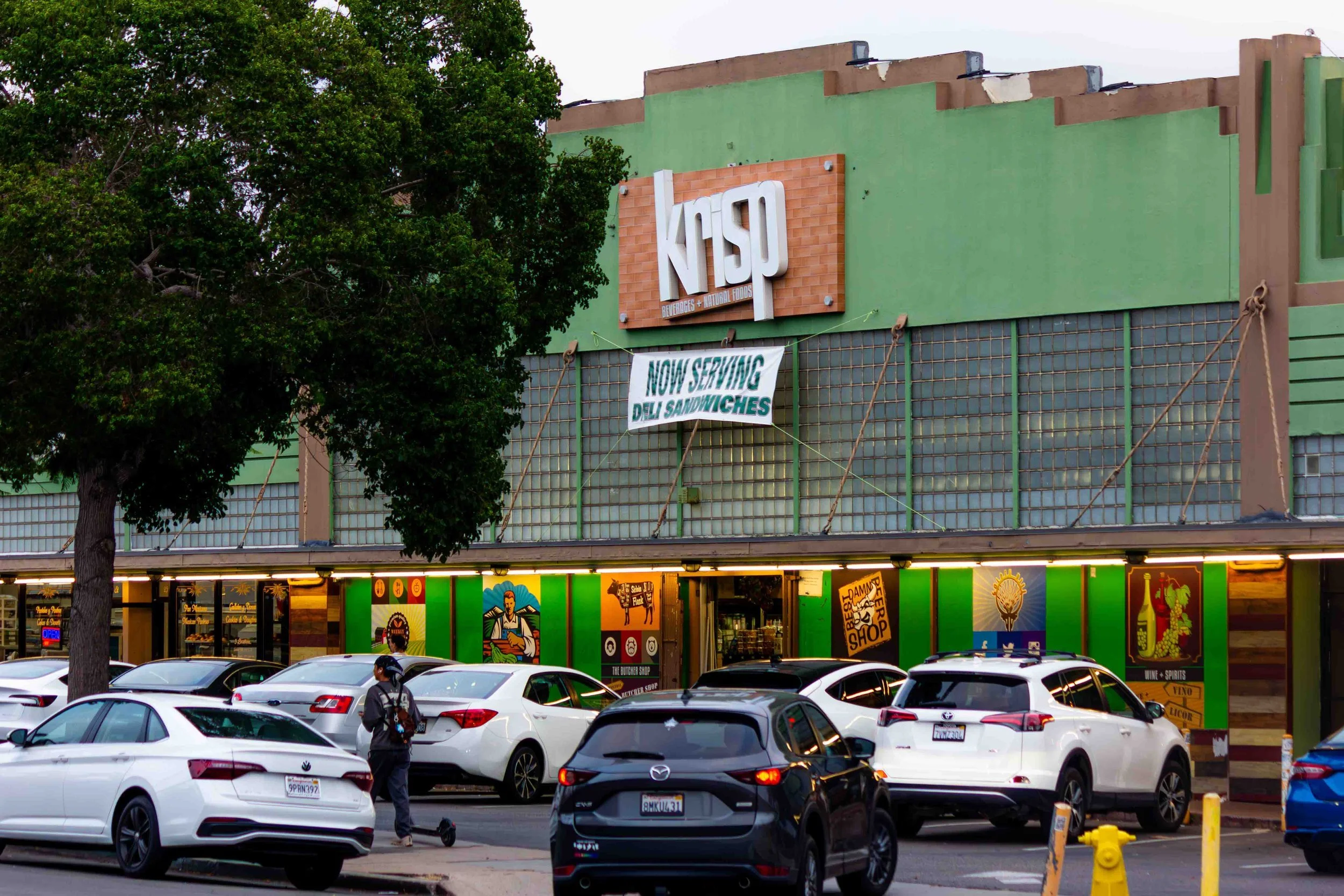 A parking lot in front of a building with a green facade, featuring a sign for Krisp and a banner announcing new deli sandwiches. Several cars are parked, and a person is standing near a white car.