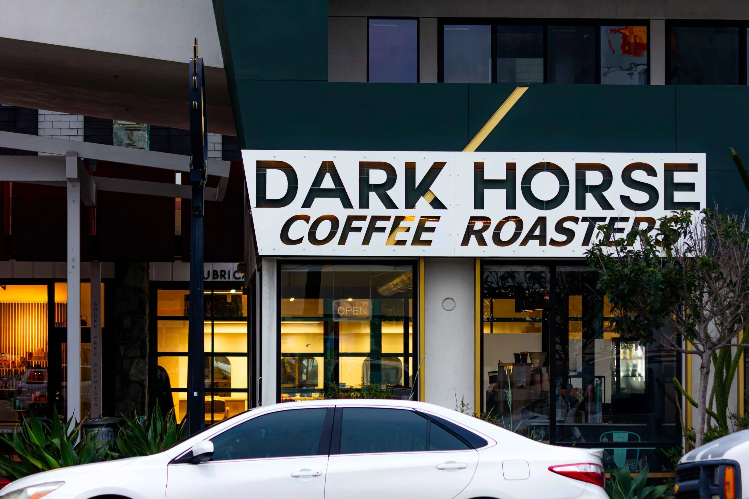 Exterior of a coffee shop called Dark Horse Coffee Roasters with a white sign displaying the name and the words 'Coffee Roasters', a large window, trees, and two cars parked in front.