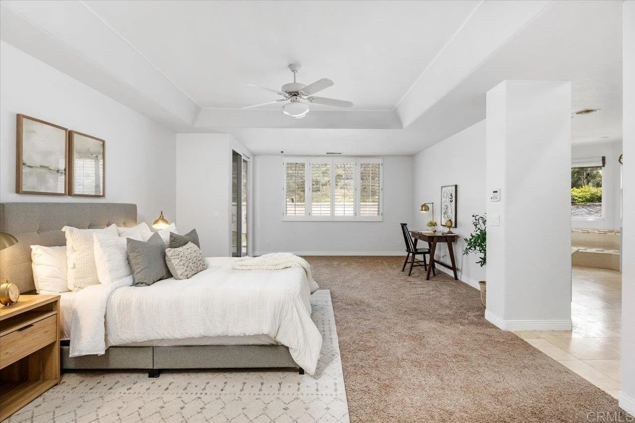 Bright, spacious bedroom with a bed, nightstand, and reading lamps, connected to a living area with window seat and desk.