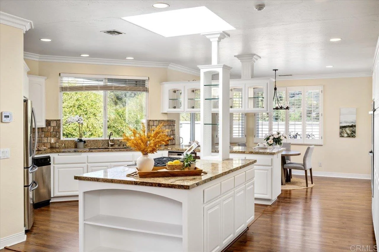 Bright kitchen with white cabinets, granite countertops, wooden flooring, a center island with a vase and fruit, and large windows with blinds, overlooking lush greenery.