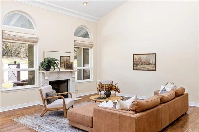 Living room with large beige sectional sofa, wooden armchair with cushion, fireplace, framed landscape pictures, large windows with Roman shades, wooden flooring, area rug, and a coffee table with decorative items.