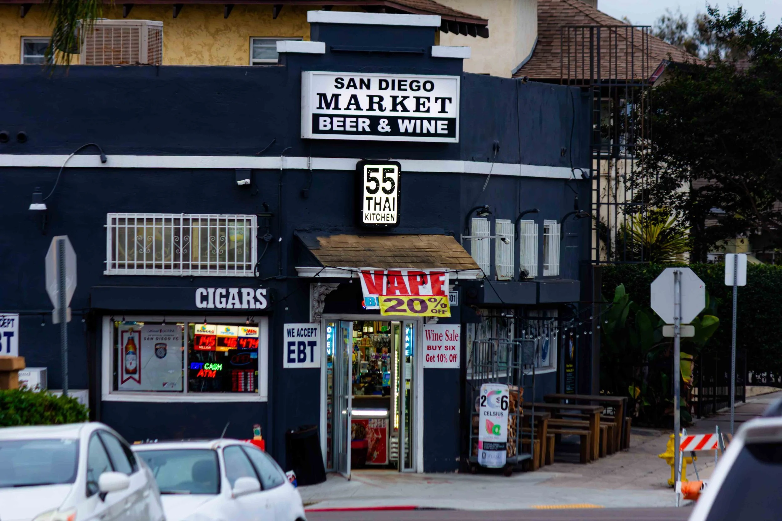 A dark-colored building with signs for a San Diego market selling beer, wine, cigars, and Thai food, with a entrance covered by a small roof and a digital sign advertising vape and a 20% discount, cars parked outside, and some outdoor seating.