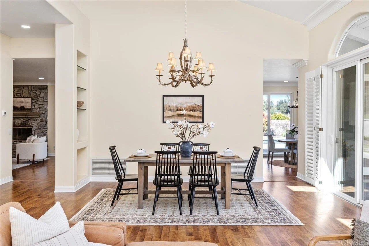 Dining room with a wooden table surrounded by six black chairs, a floral centerpiece in a blue vase, and a chandelier hanging above. There is a picture on the wall and a rug underneath the table. Sunlight streams through large glass doors with white 