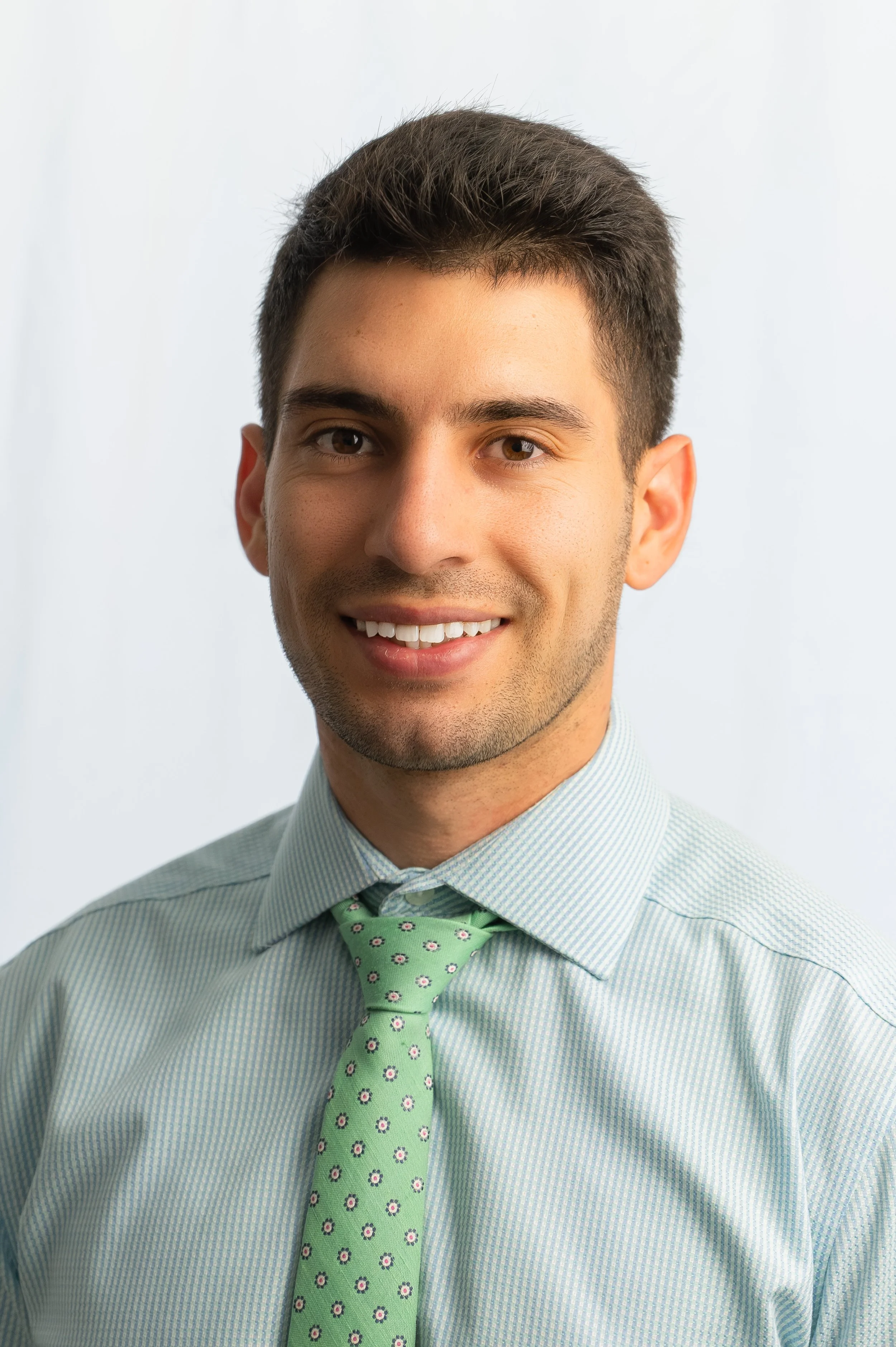 Headshot of a young man with dark hair wearing a light blue dress shirt and a green tie with small floral patterns, smiling at the camera.