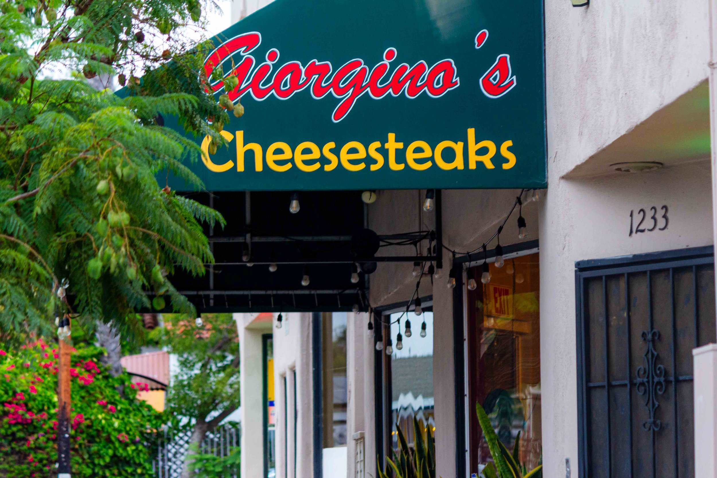 Sign for Giorgio's Cheesesteaks restaurant on a building exterior, with string lights hanging below. There is greenery and flowers nearby.