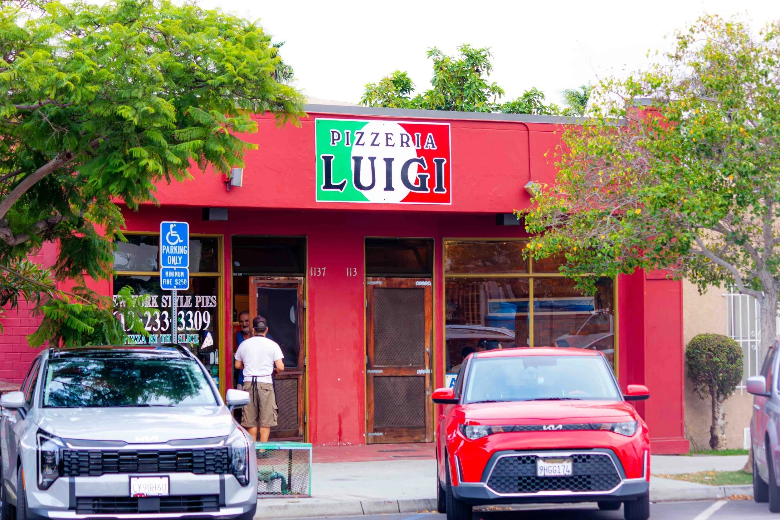 A red building with a sign that reads "Pizzeria Luigi" and a menu board outside. There are cars parked in front and people entering the restaurant.