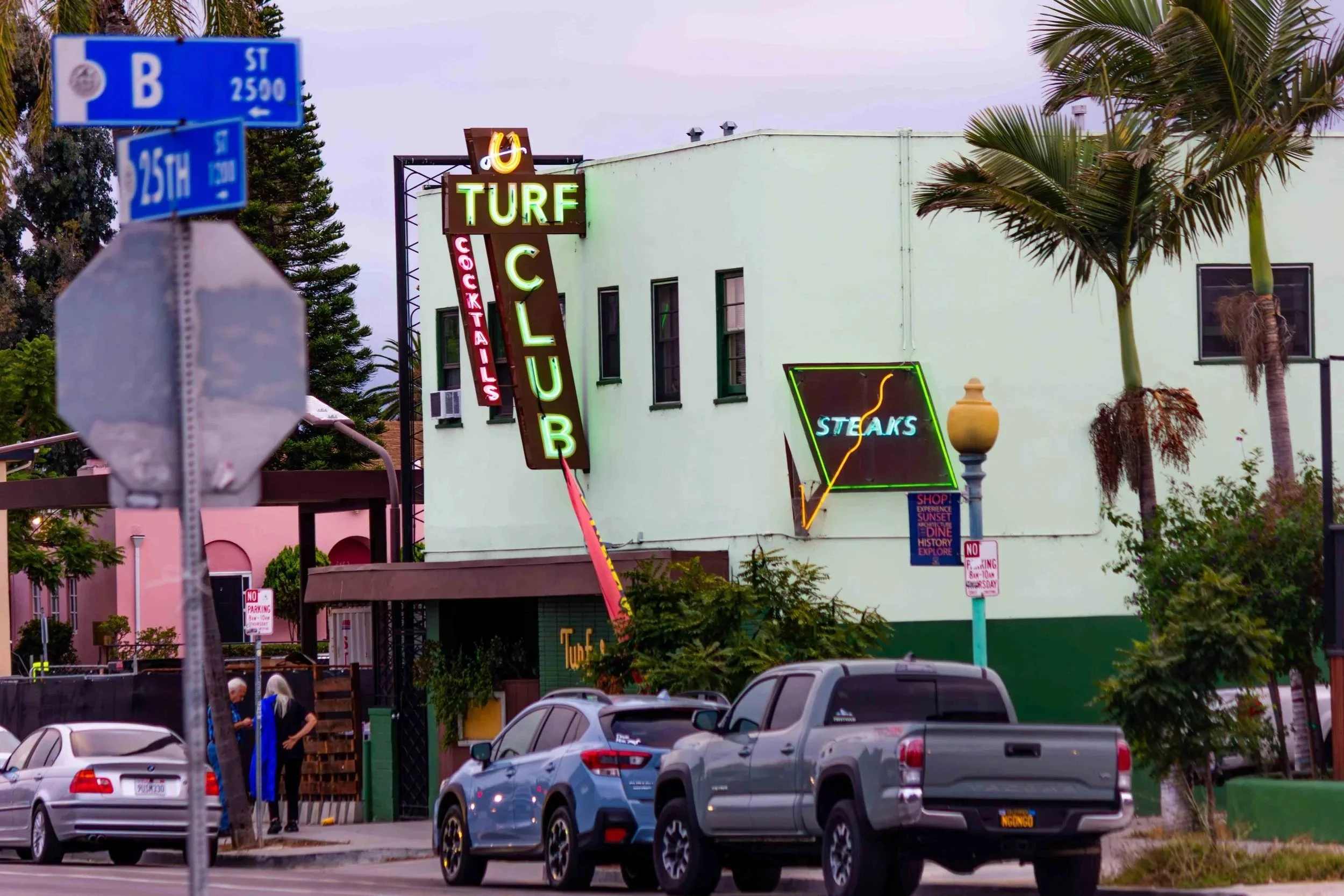 Street scene featuring a building with signs for a club, steakhouse, and cocktails, with palm trees, parked cars, and a street sign for 25th Street in the foreground.