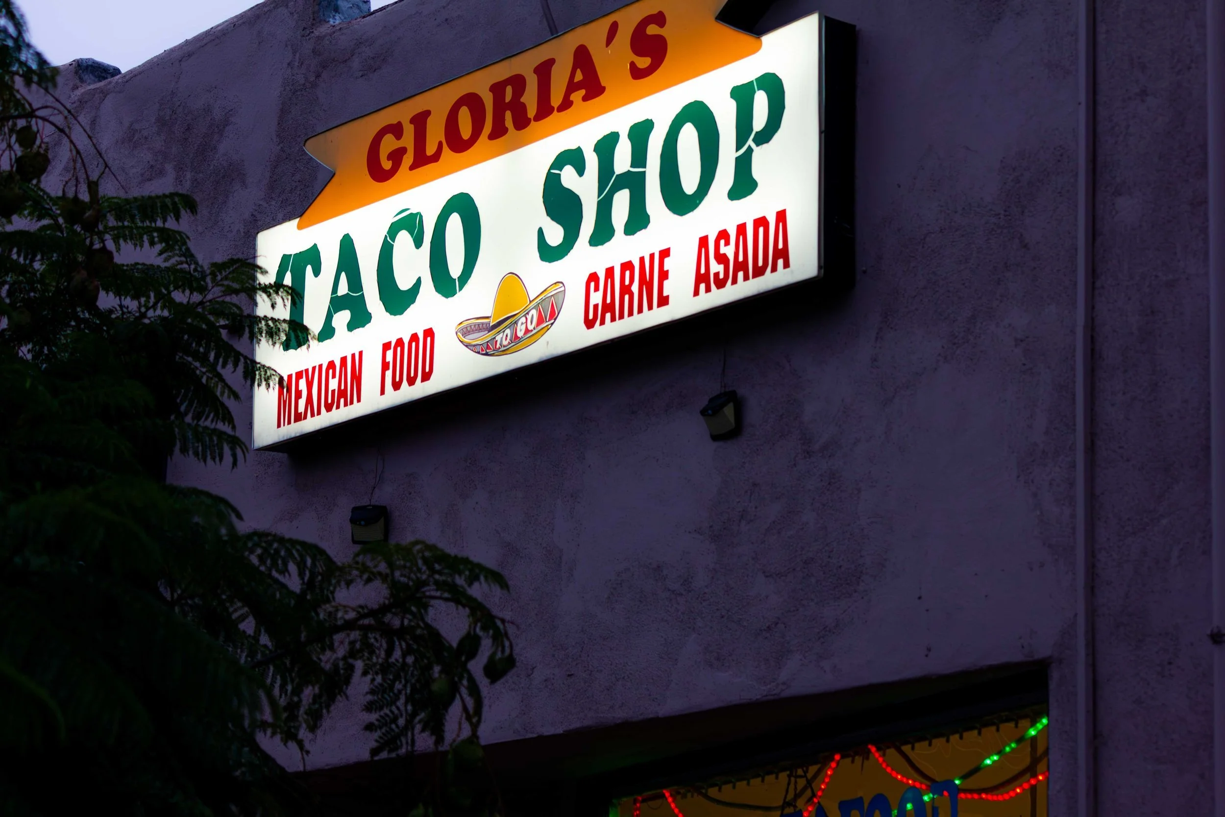  illuminated sign for Gloria's Taco Shop advertising Mexican food and carne asada, with a decorative sombrero logo, mounted on a stucco wall exterior, partially obscured by tree branches.