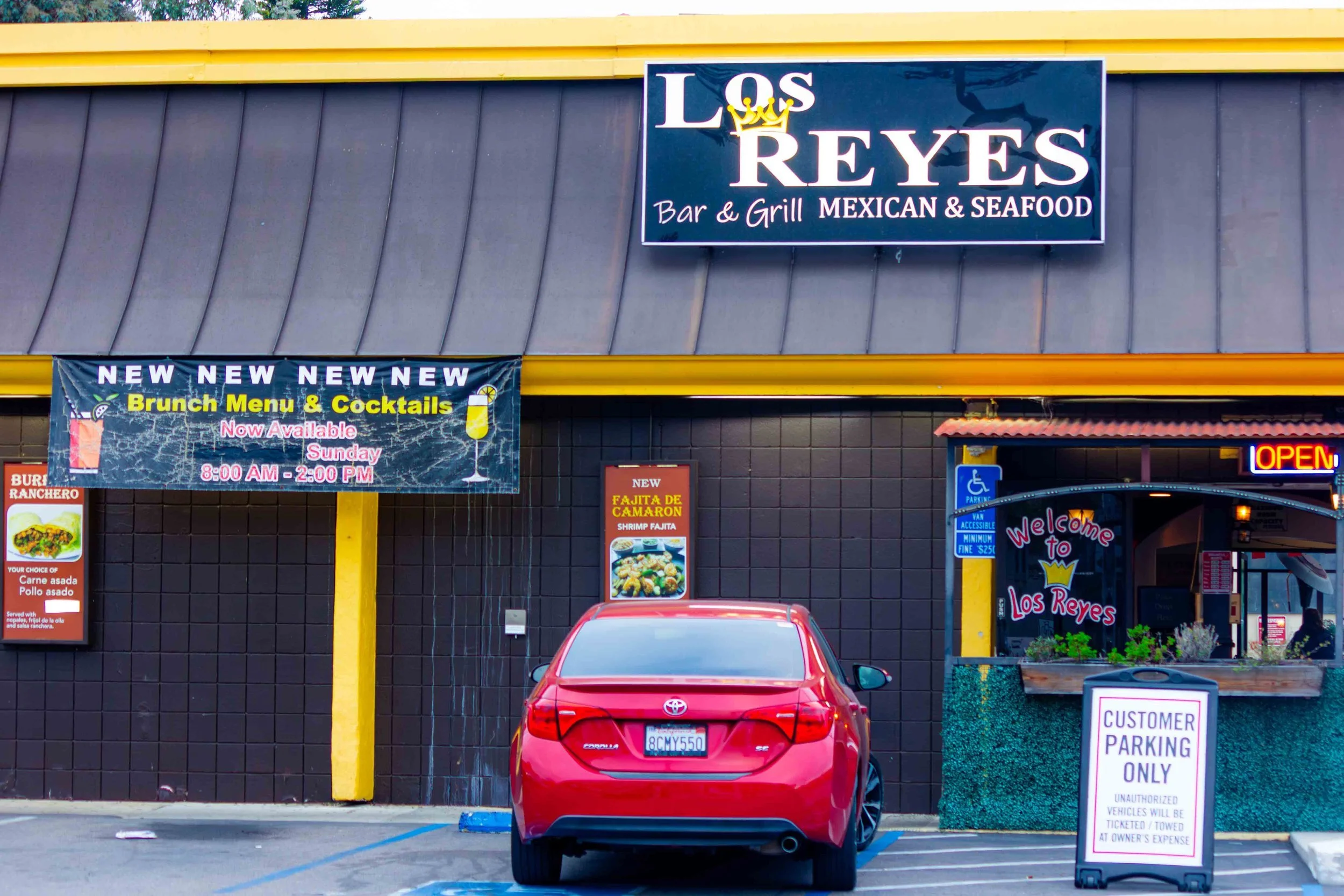 Front of a Mexican restaurant called Los Reyes with a red car parked in front. The restaurant has a black and yellow exterior, a large sign on top, and advertisements for brunch and new menu items, including shrimp fajitas. A welcome sign and a custo