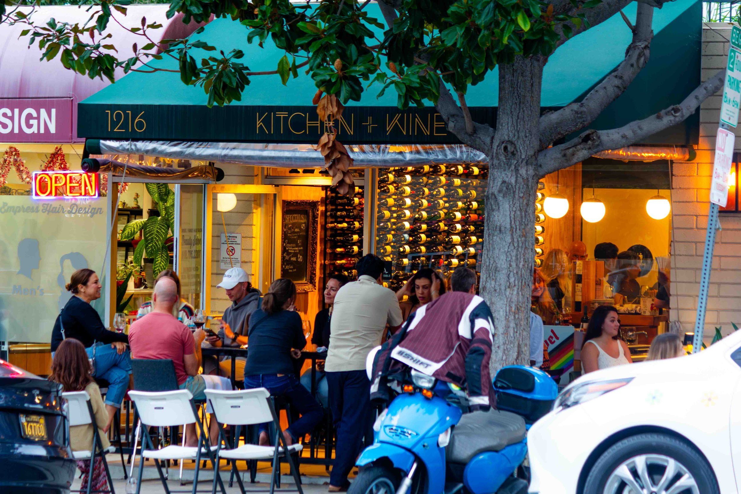 A lively outdoor dining scene in front of a restaurant named 'Kitchen + Wine' with a crowd of people seated at tables, a tree, a parked scooter, and cars on the street.