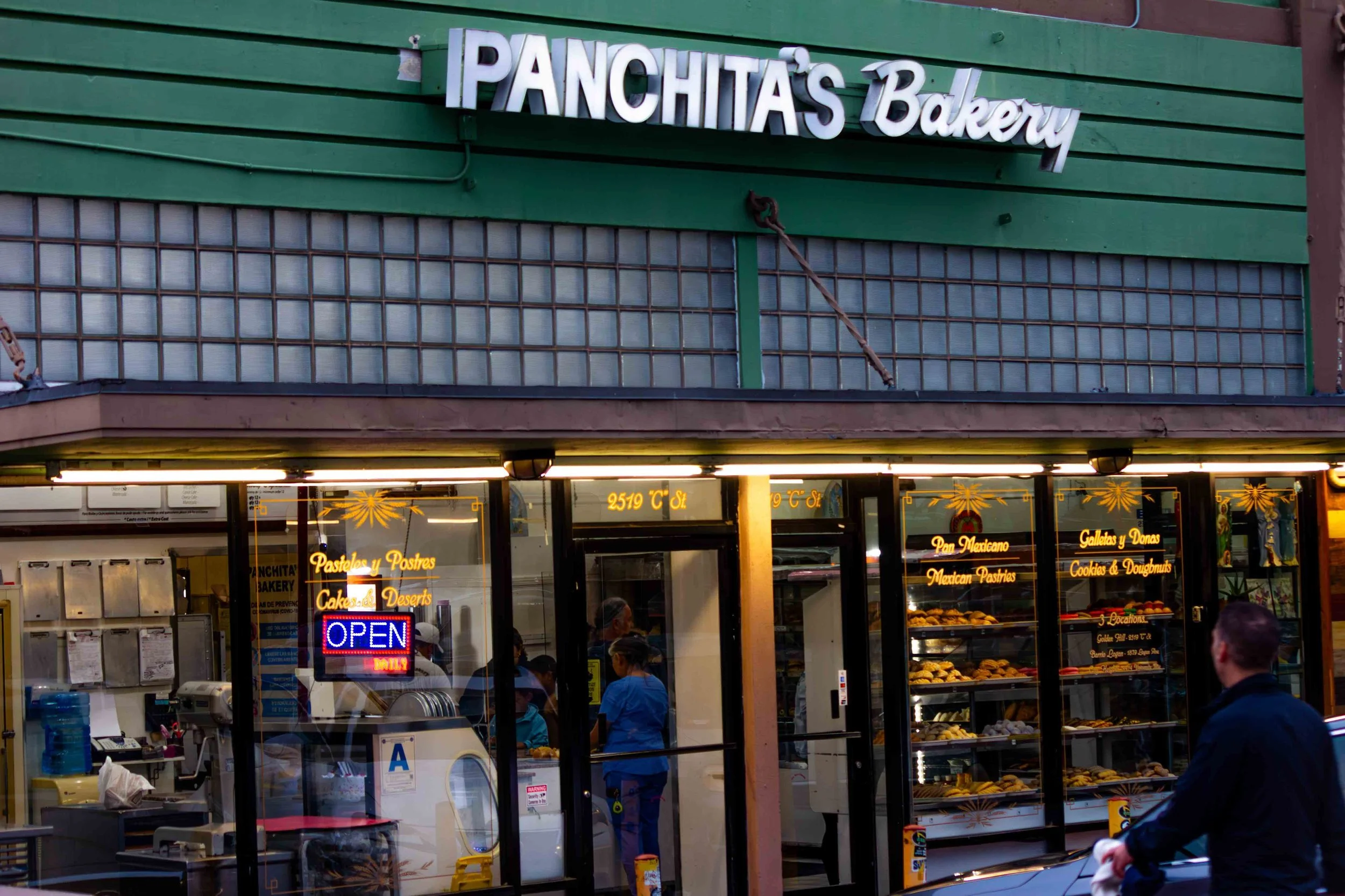 Exterior of Panchita's Bakery with glass display case showing baked goods, neon 'Open' sign, and customers inside and outside the shop.