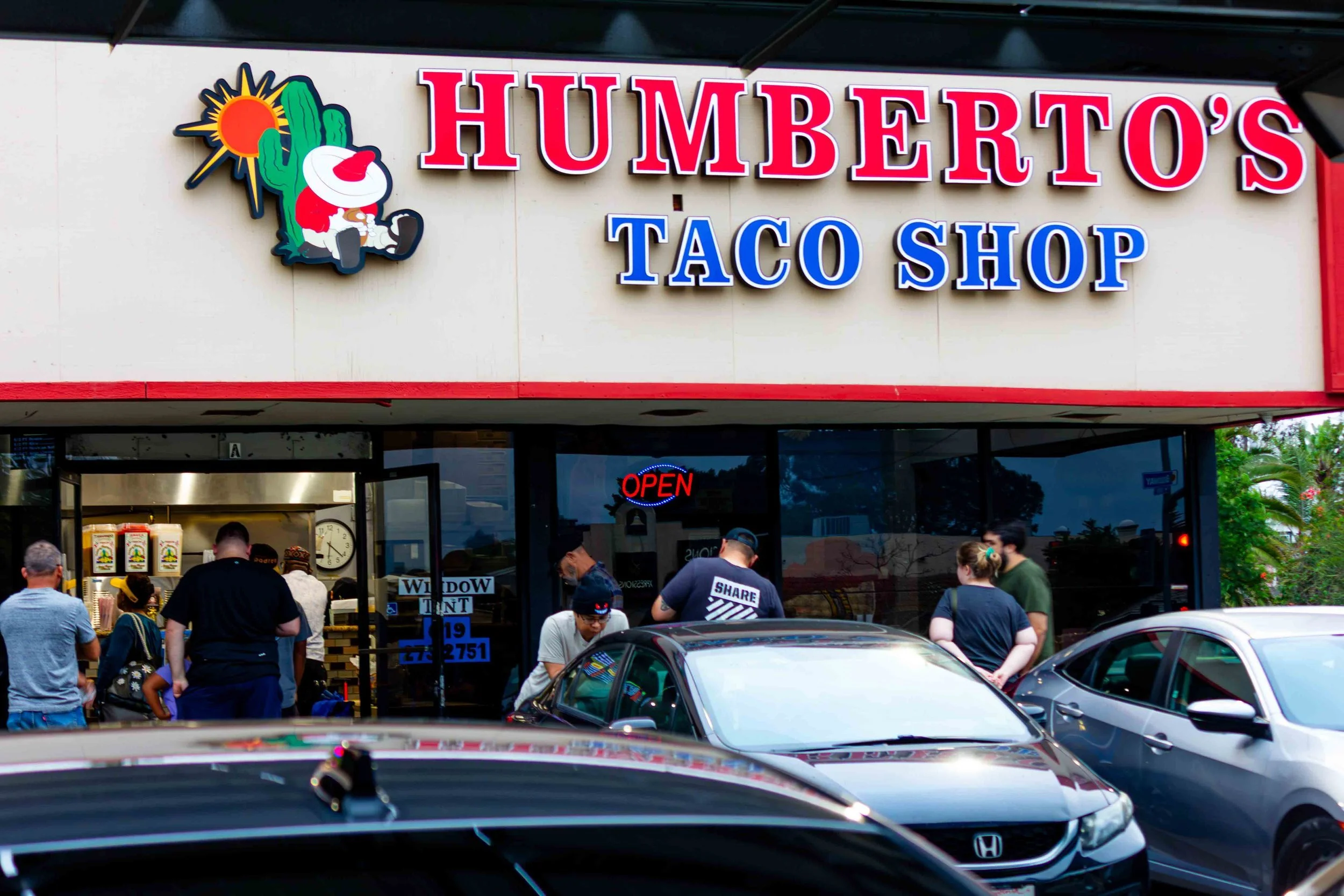 Humberto's Taco Shop storefront with people standing outside and cars parked in front.