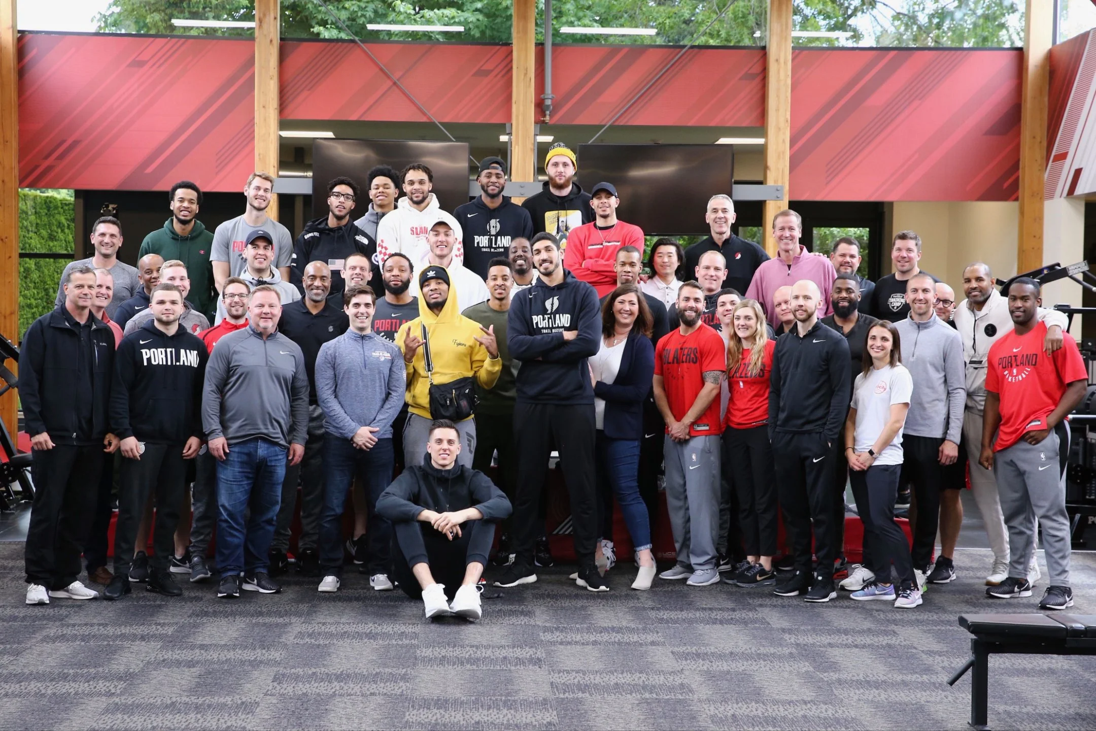Group of approximately 35 people, including men and women of various ages and ethnicities, pose together inside a building with large windows and wooden beams. Many are wearing athletic or casual clothing, some with Portland Trail Blazers apparel. They are smiling and standing close together, with a few making hand gestures.