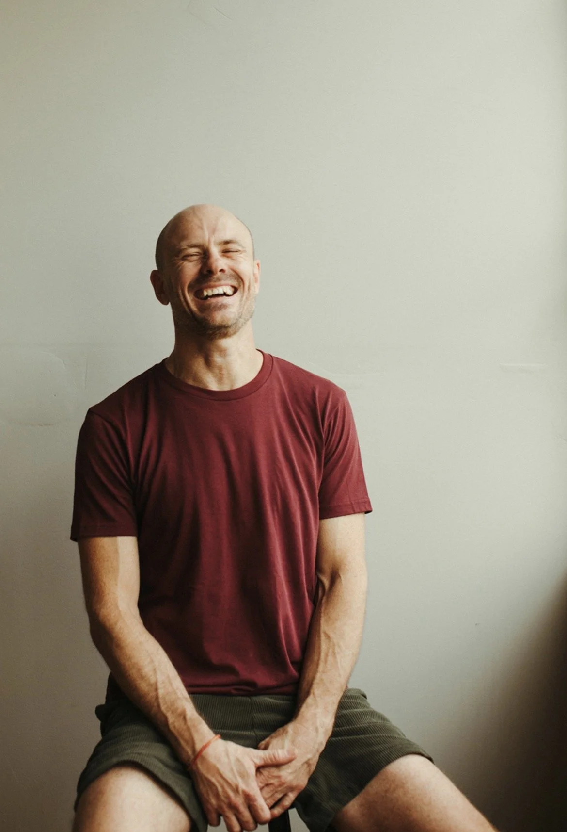 A man sitting and laughing against a plain light-colored wall, wearing a maroon t-shirt and shorts.