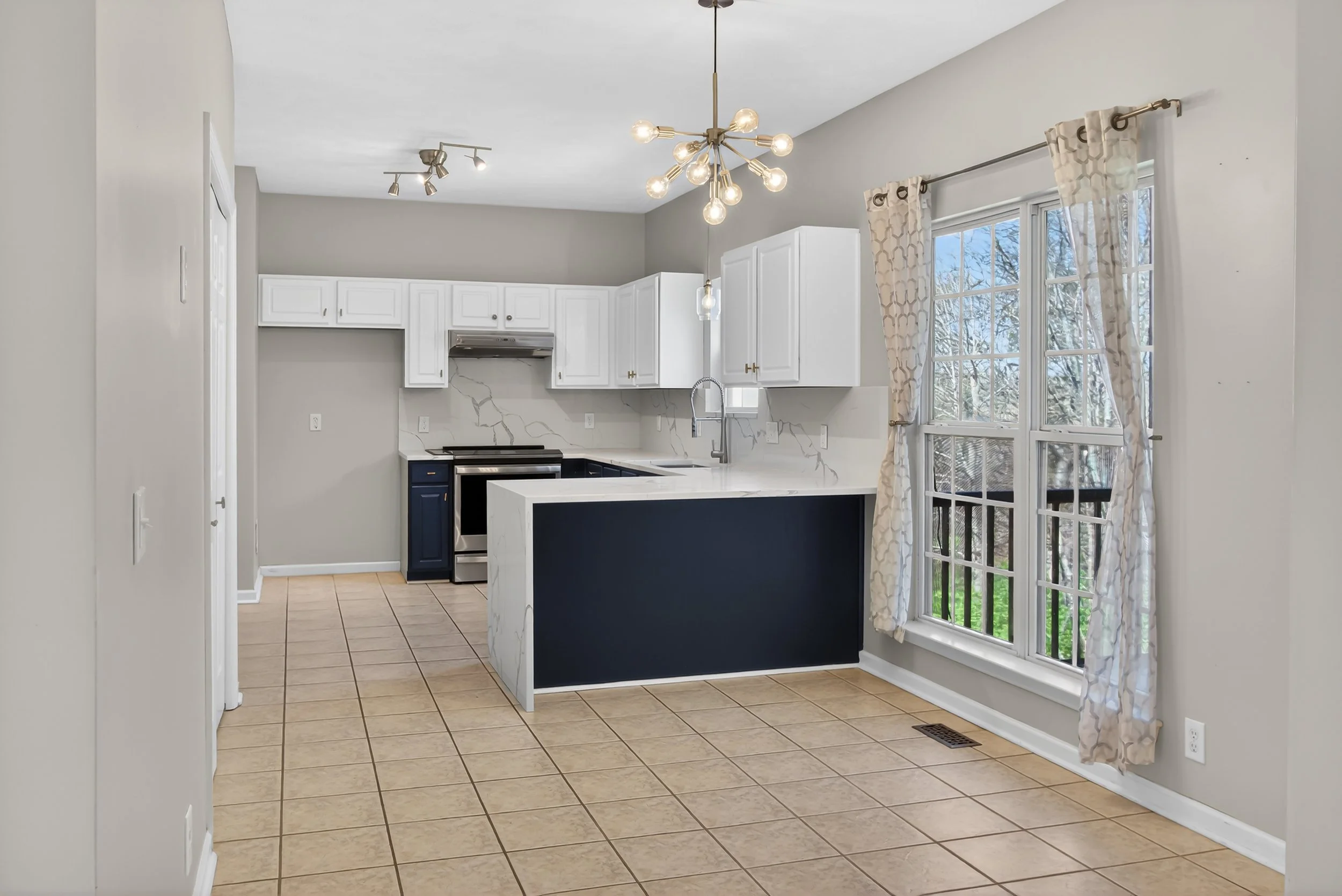 Modern kitchen with white cabinets, a marble countertop, a stainless steel stove, and a large window with patterned curtains, illuminated by contemporary light fixtures.