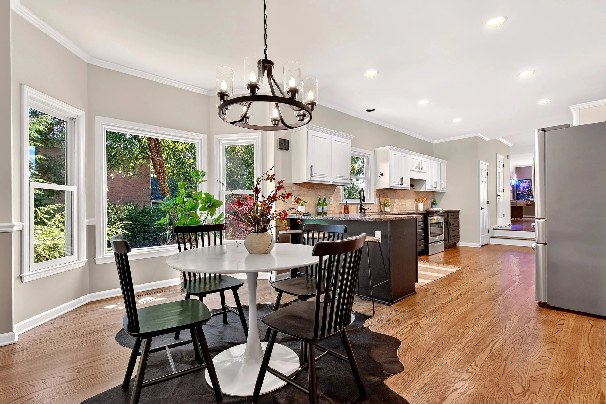 Bright kitchen with white cabinets, large windows overlooking greenery, black dining chairs around a white round table with a plant centerpiece, and a black chandelier overhead.
