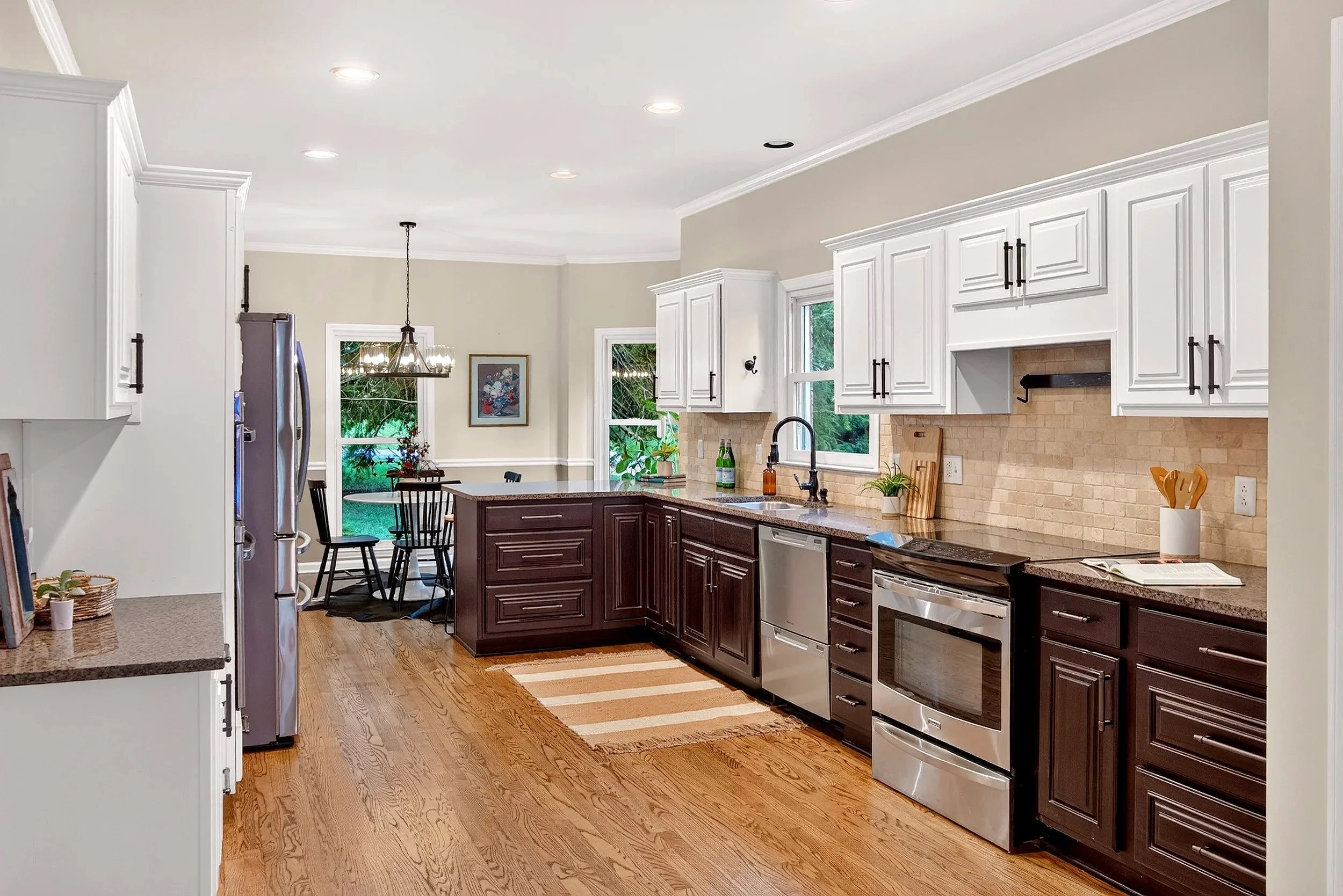 A spacious kitchen with white upper cabinets, dark lower cabinets, a granite countertop, and stainless steel appliances. There is a dining area with a table and chairs visible through the doorway, windows letting in natural light, and a hardwood floo