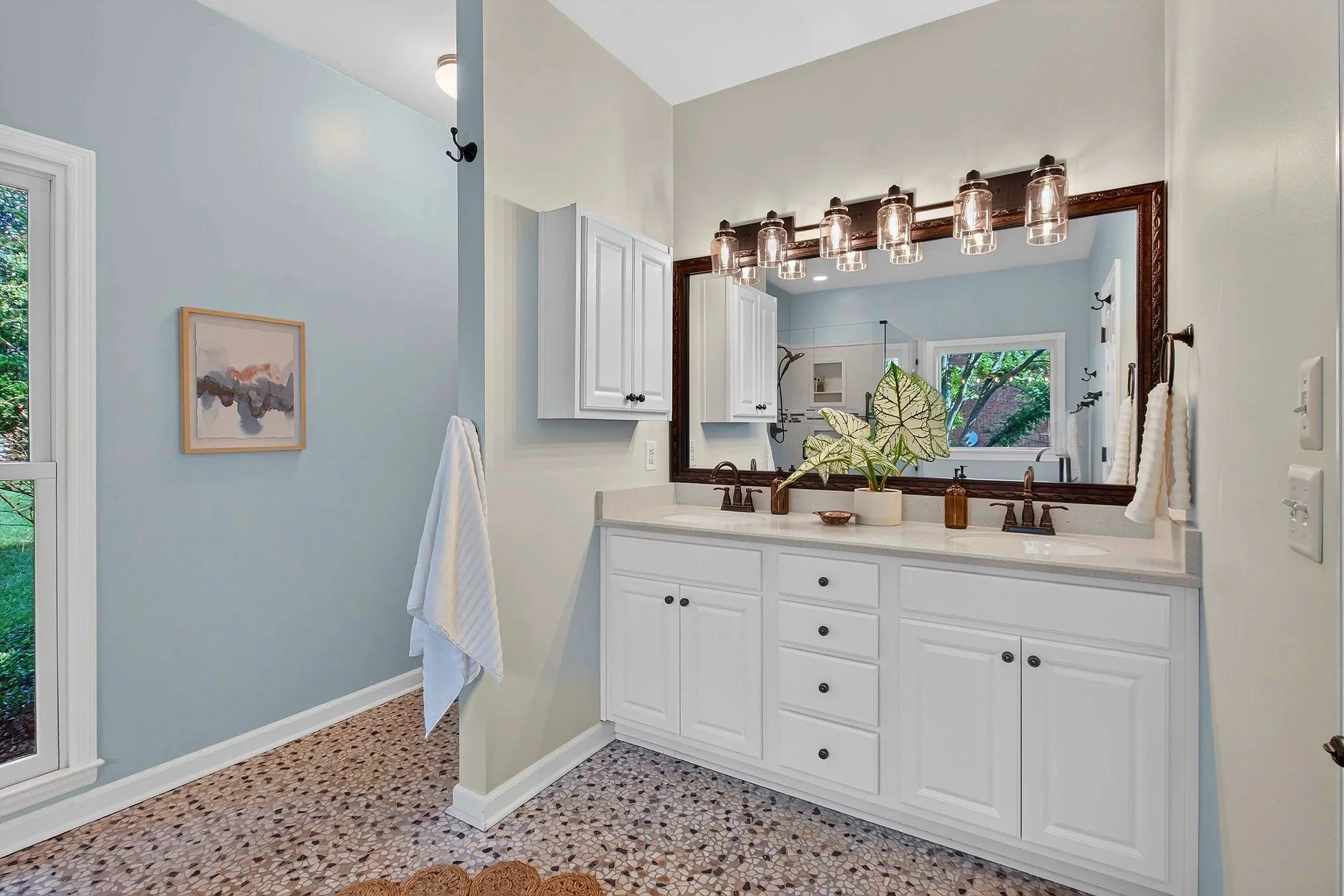 Bathroom with light blue walls, white cabinets, large mirror, vanity with potted plant and toiletries, window showing trees outside, and mosaic tile floor.