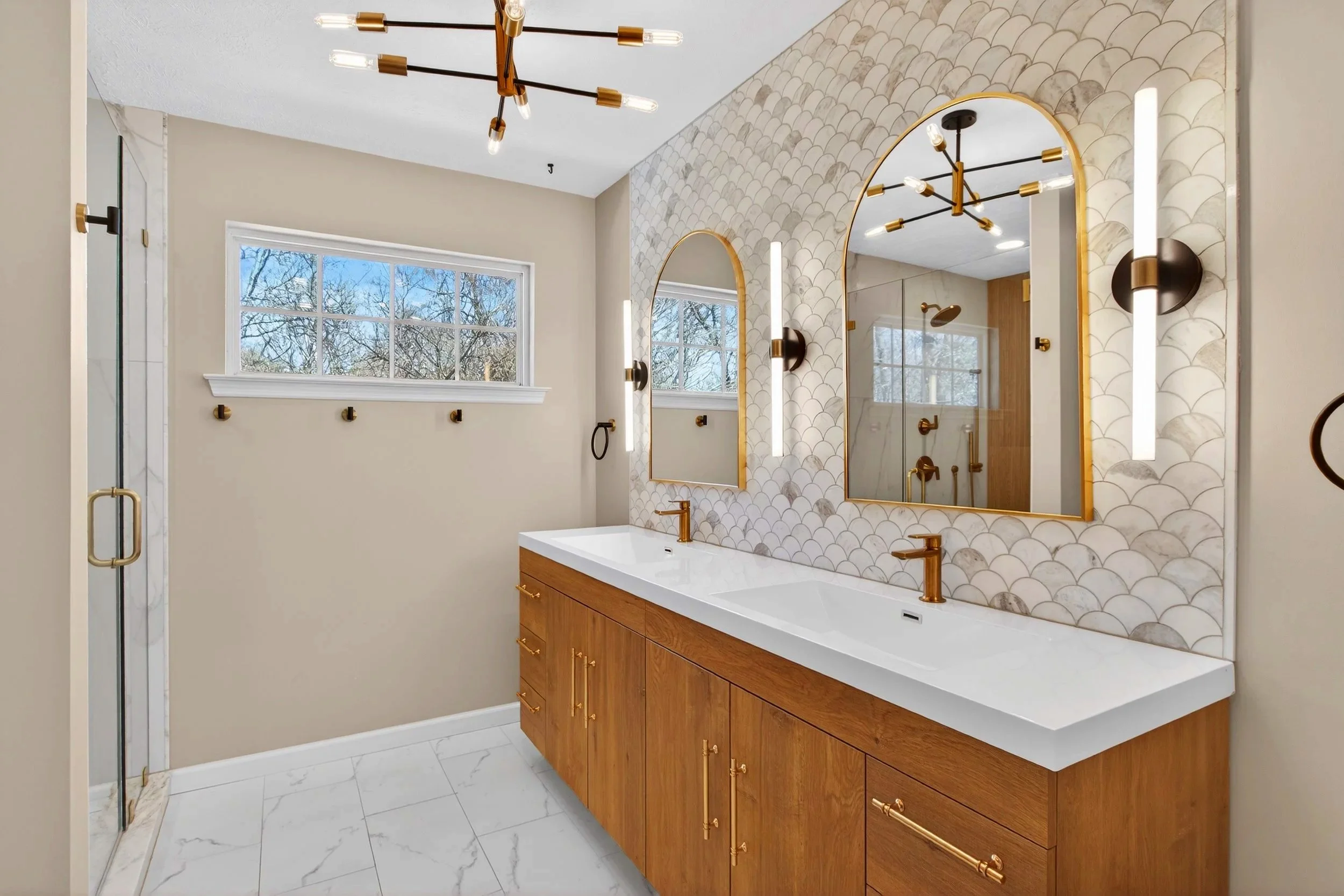 Modern bathroom with a double vanity featuring wooden cabinets, white countertop, and gold fixtures. There are two large arched mirrors with gold frames, black wall sconces, and a natural stone tile backsplash. A window with a view of trees is above 