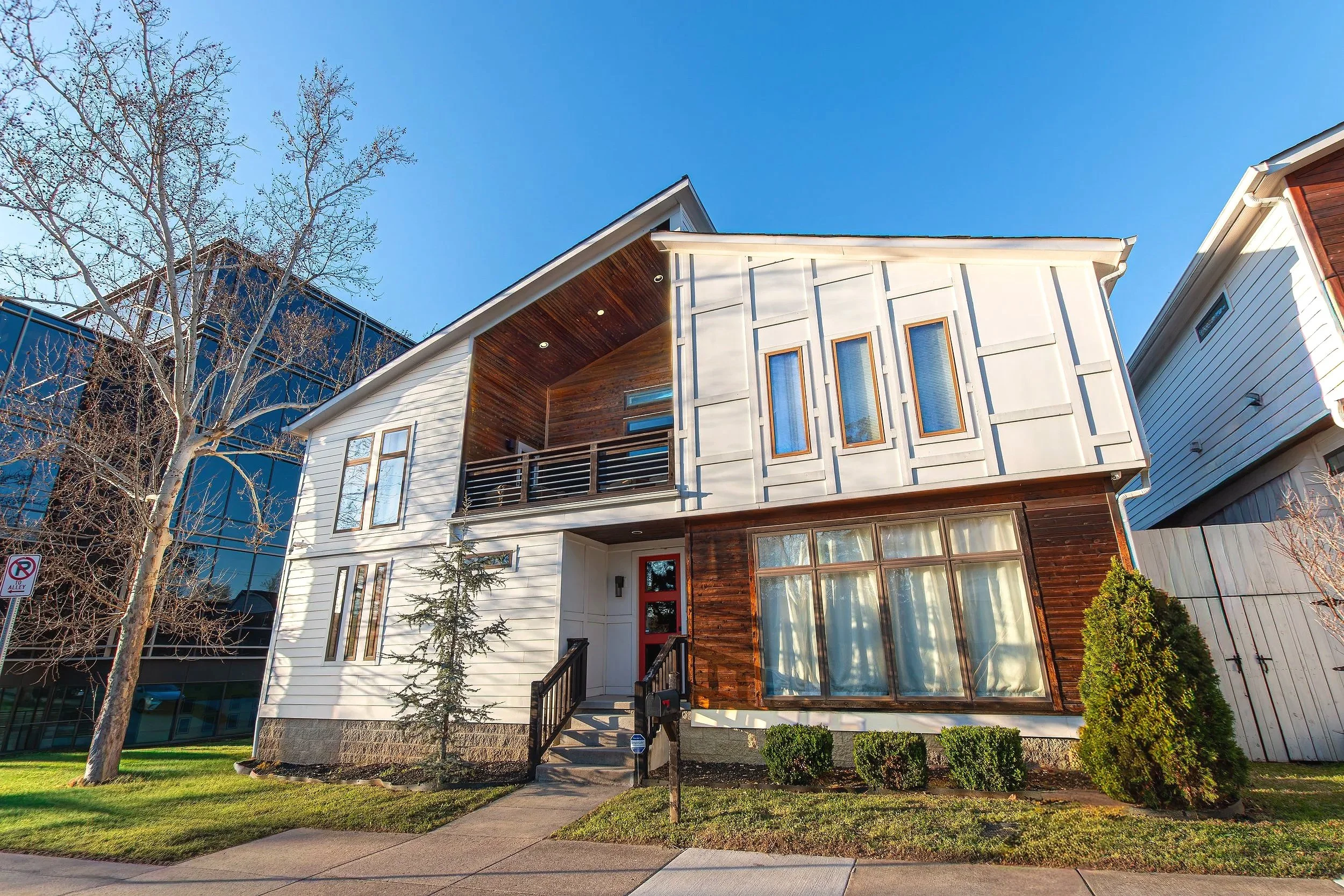 A modern two-story house with white siding, large windows, and a wooden front door, surrounded by small bushes and a green lawn, with leafless trees and a blue sky in the background.