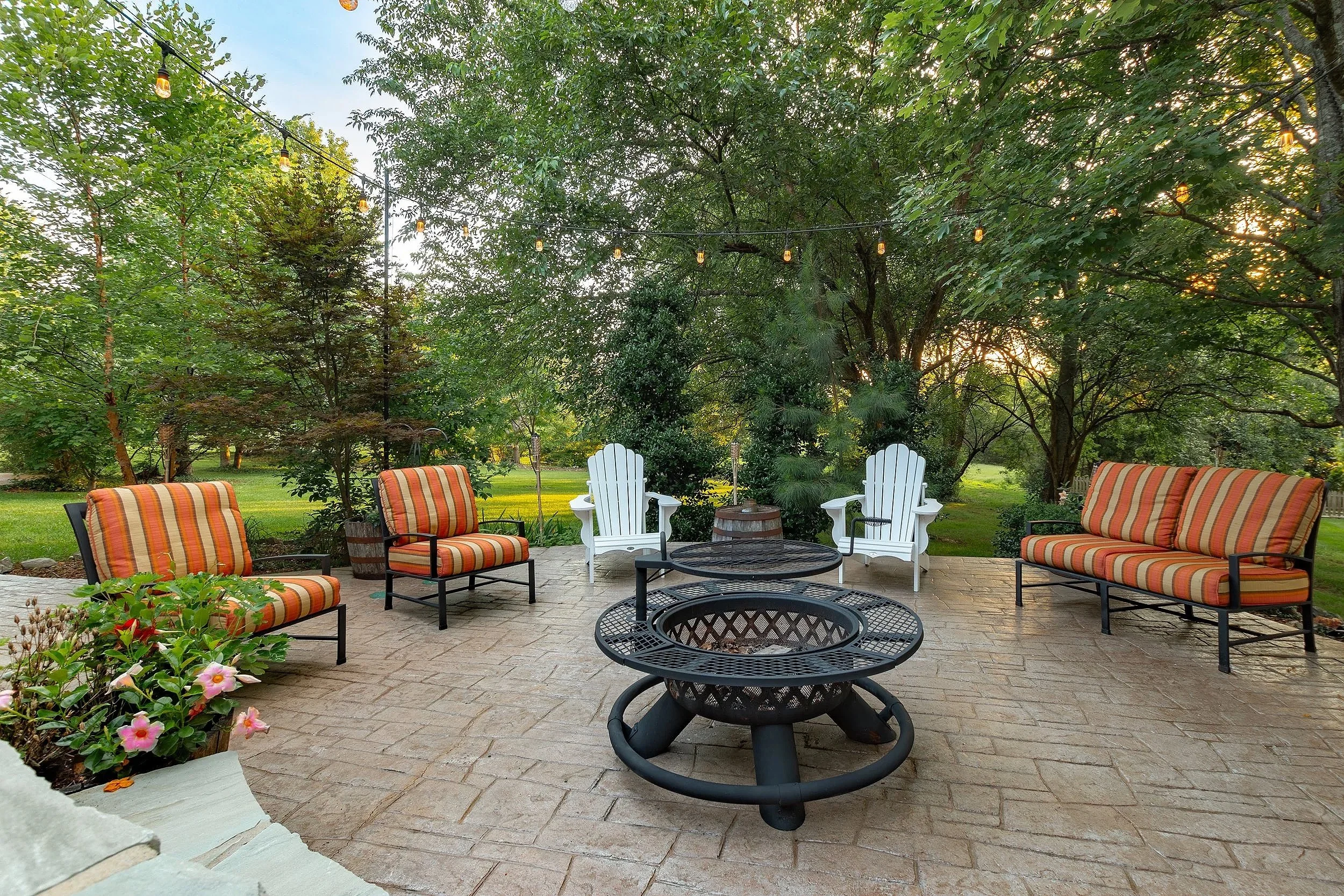 A backyard patio with outdoor seating, including striped cushioned chairs around a fire pit, surrounded by lush green trees and string lights, with the sun setting in the background.