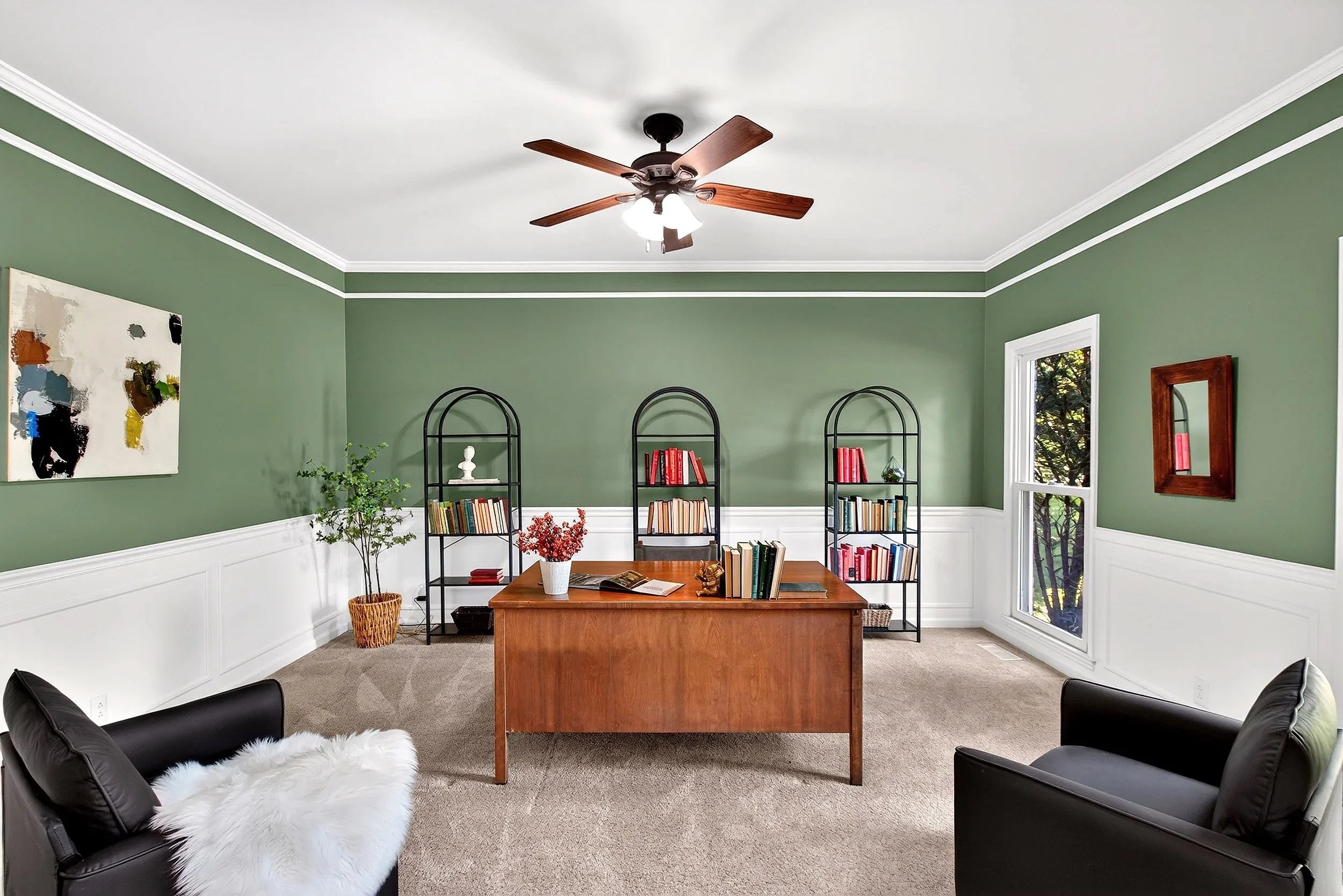 Heated living room with dark leather chairs, a wooden coffee table, green walls, white trim, a ceiling fan, and black bookshelf with red and wooden books, a window showing trees, and abstract art on the wall.