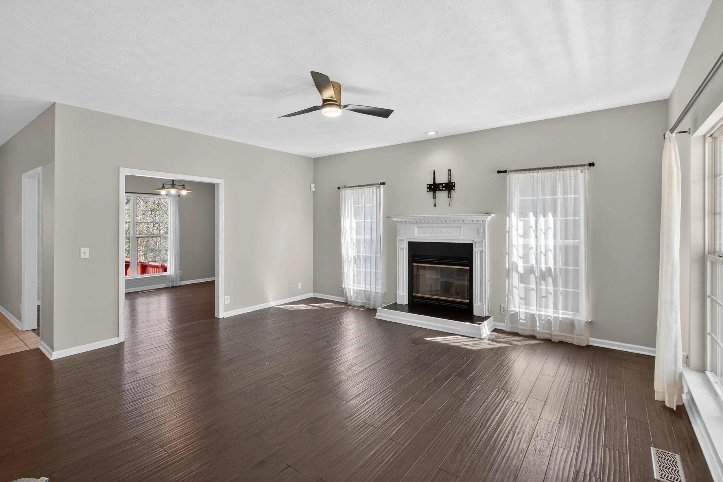 Living room with dark wood floors, white walls, a fireplace with decorative trim, and large windows with sheer curtains. A ceiling fan and recessed lighting are also visible.