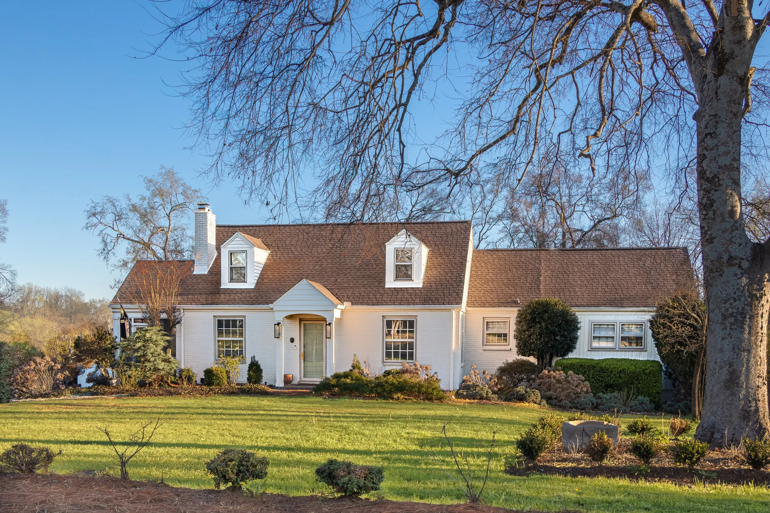 A white house with a brown shingle roof, dormer windows, and a front porch, surrounded by a well-maintained yard with bushes and small trees. There are large leafless trees in the background under a clear blue sky.