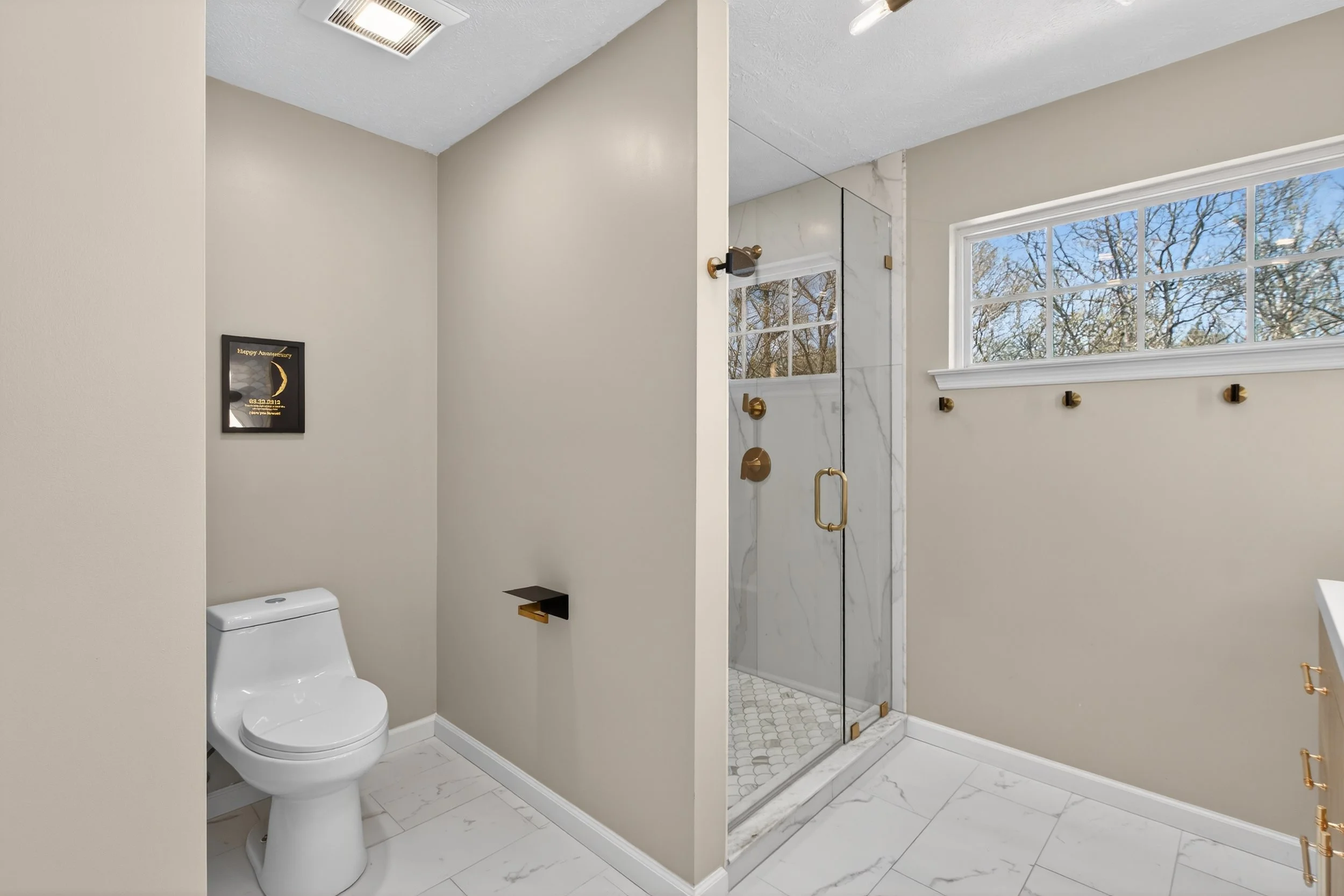 A modern bathroom featuring a white toilet, a glass-enclosed shower with gold fixtures, a large window with a view of trees, and minimal decor.