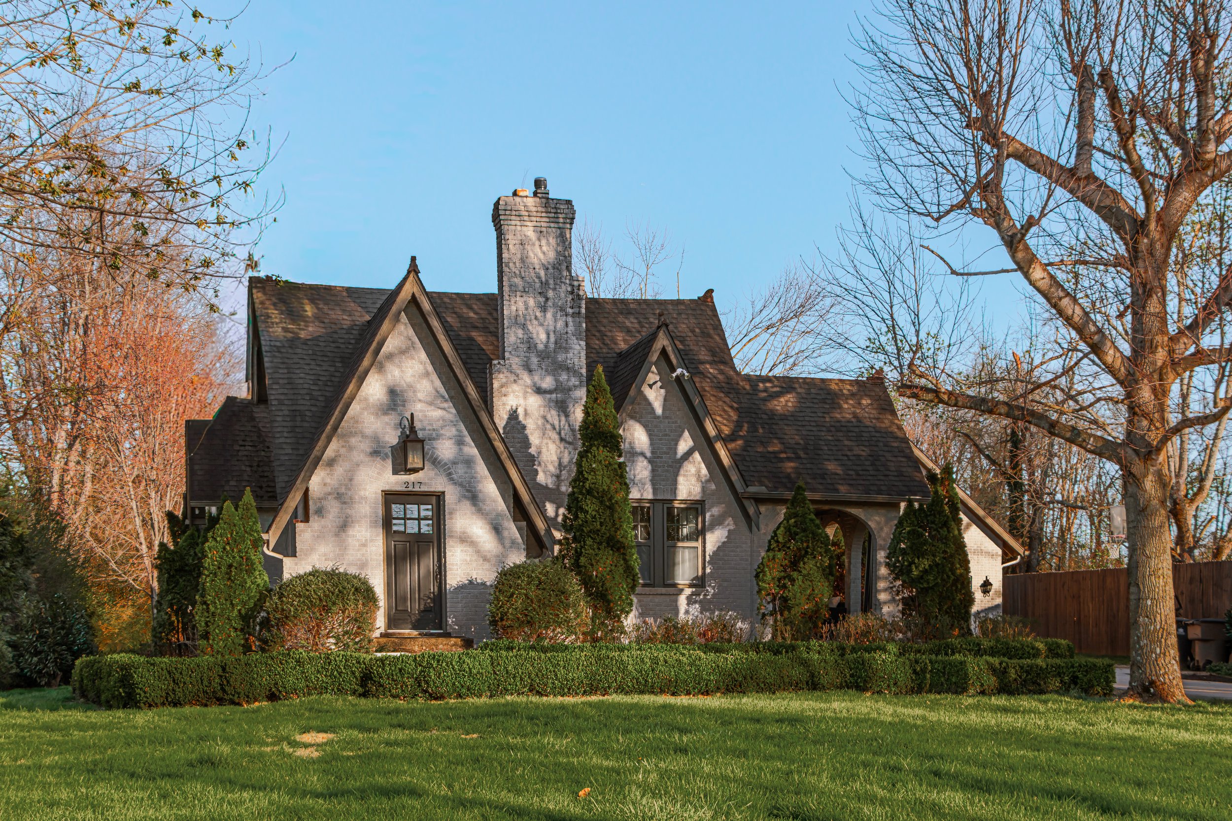 A house with a steep gabled roof, white brick exterior, and a chimney. The house is surrounded by trees and bushes, with well-maintained lawn in front, during daytime with clear blue sky.