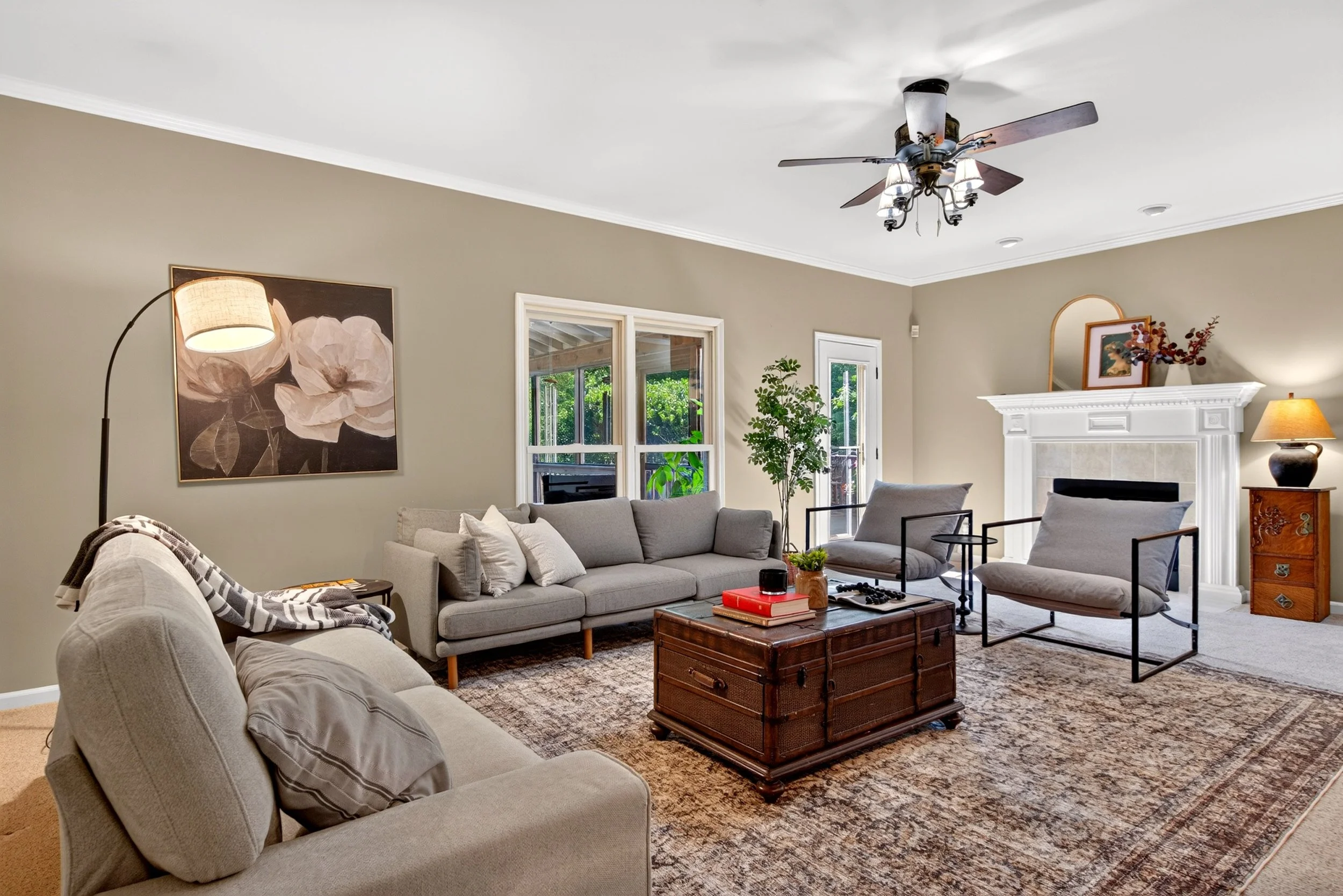 Living room with beige walls, large windows, and a white fireplace mantel. Furnished with a gray sofa, black metal and beige armchairs, a wooden chest coffee table, and various decorative items including a painting of flowers, a table lamp, and green