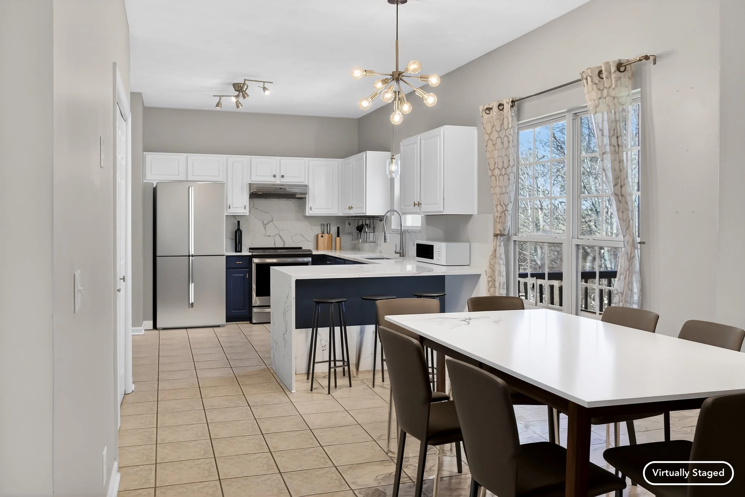 Kitchen with white cabinets, stainless steel refrigerator, marble island, dining table with dark chairs, large window with patterned curtains, and unique ceiling light fixtures.