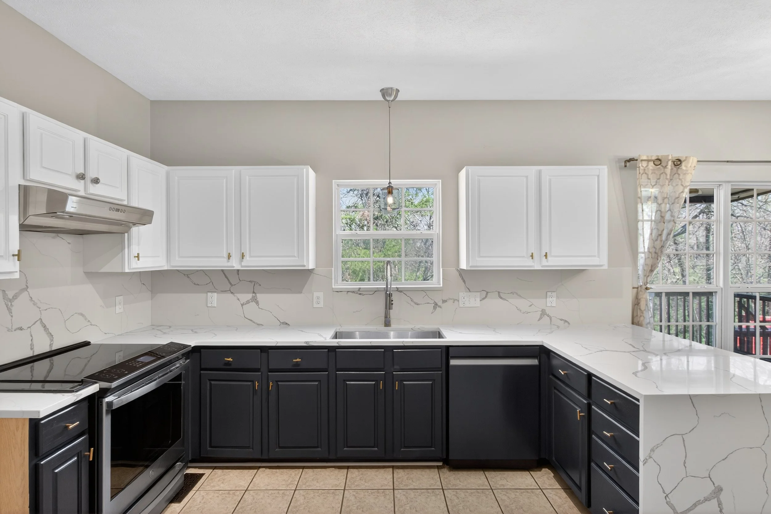 Modern kitchen with white upper cabinets, dark lower cabinets, marble countertops, a stove, and windows with curtains, overlooking trees.