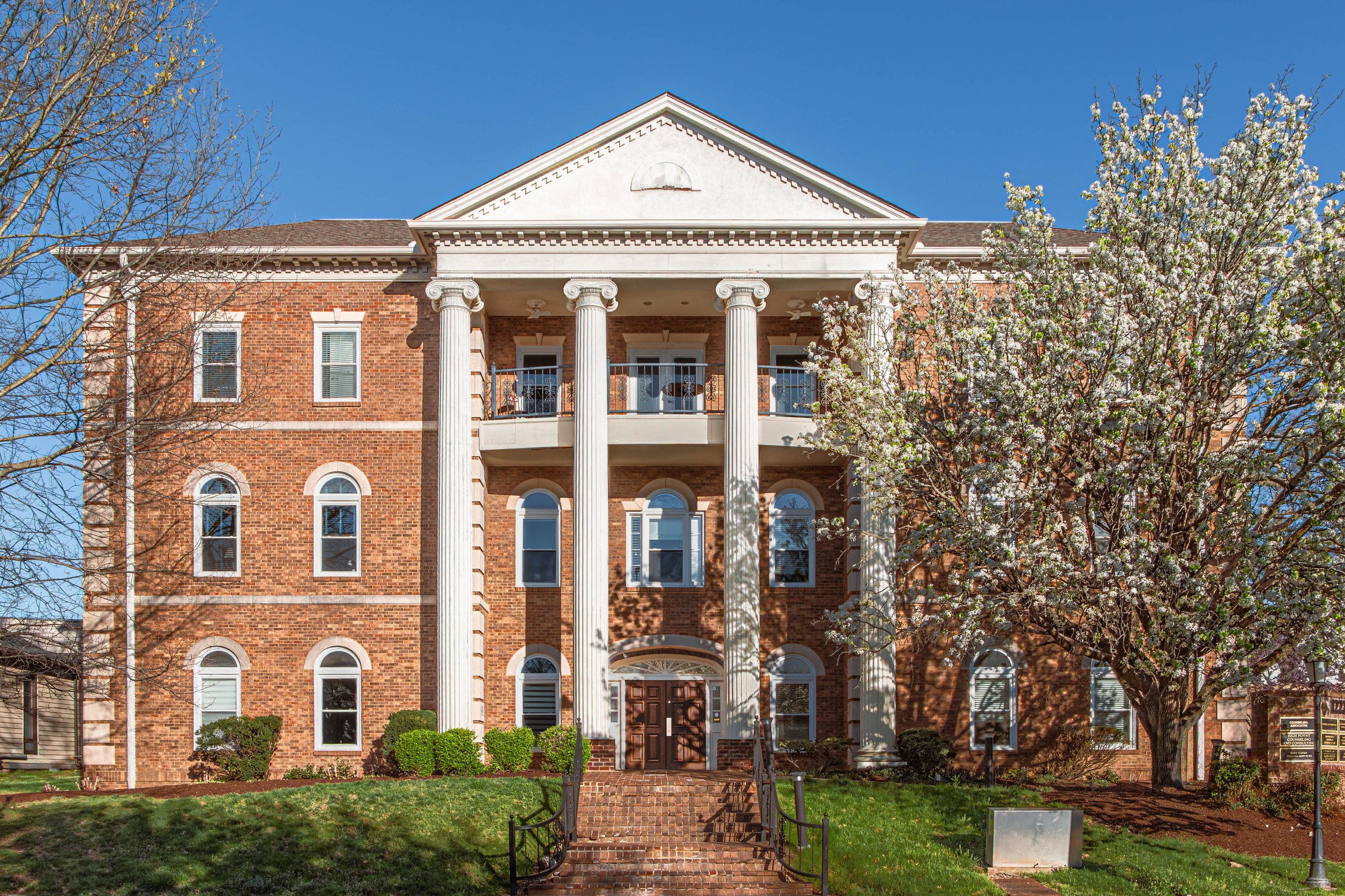 A red brick building with white columns and a porch, surrounded by trees and greenery, under a clear blue sky.