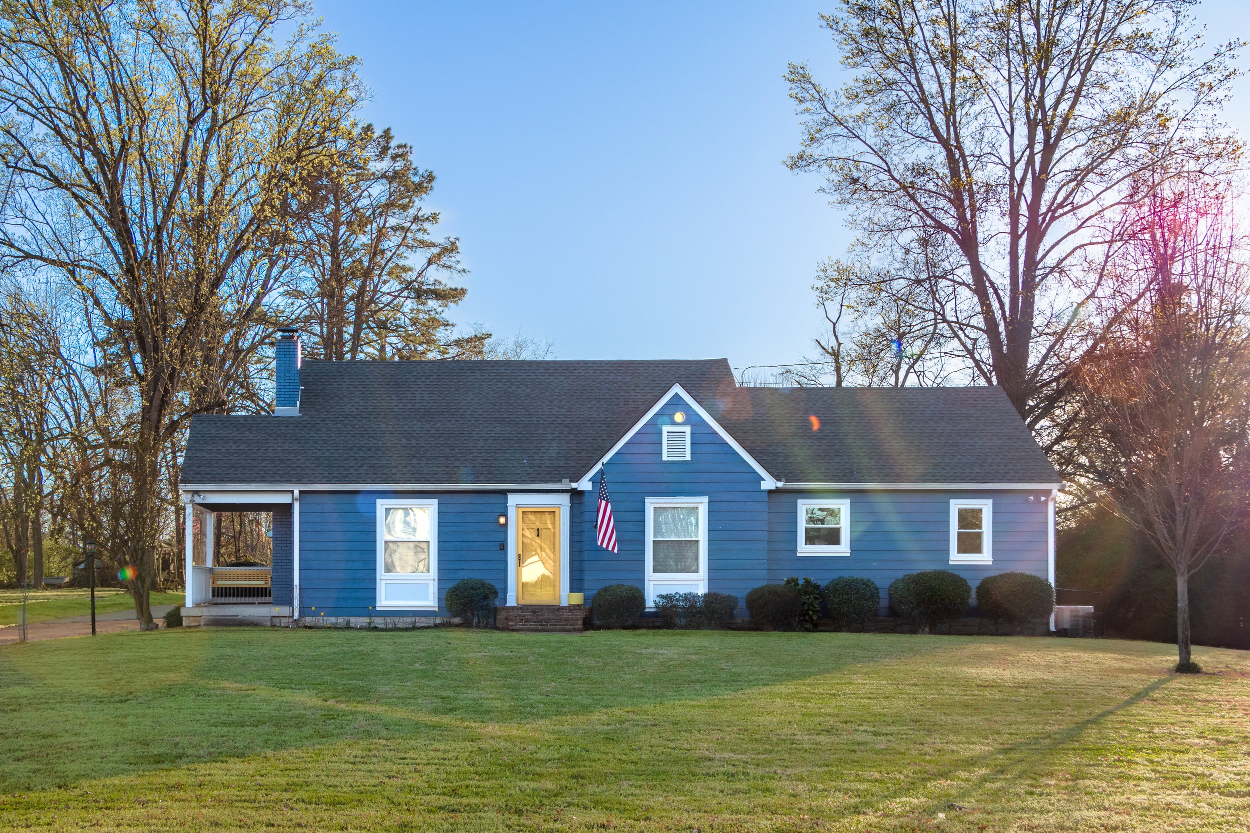 A blue house with a dark gray roof and white trim, front yard with green grass, small bushes, and trees with budding leaves, under a clear blue sky with the sun shining and creating lens flare.