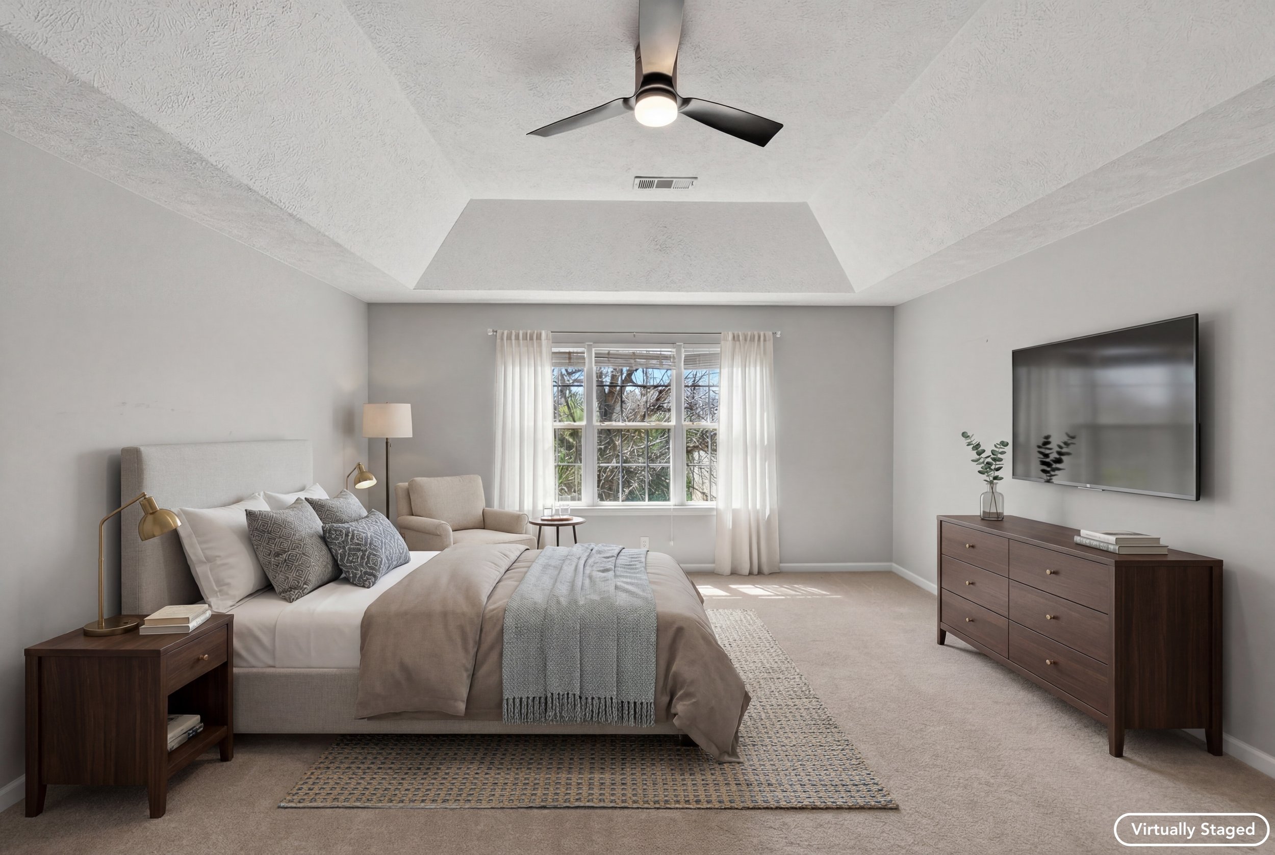 Bright, cozy bedroom with a large window, white curtains, and a bed with neutral bedding and decorative pillows. A wooden nightstand with a lamp and books is beside the bed. There is a wooden dresser with a plant and books. A flat-screen TV is mounte